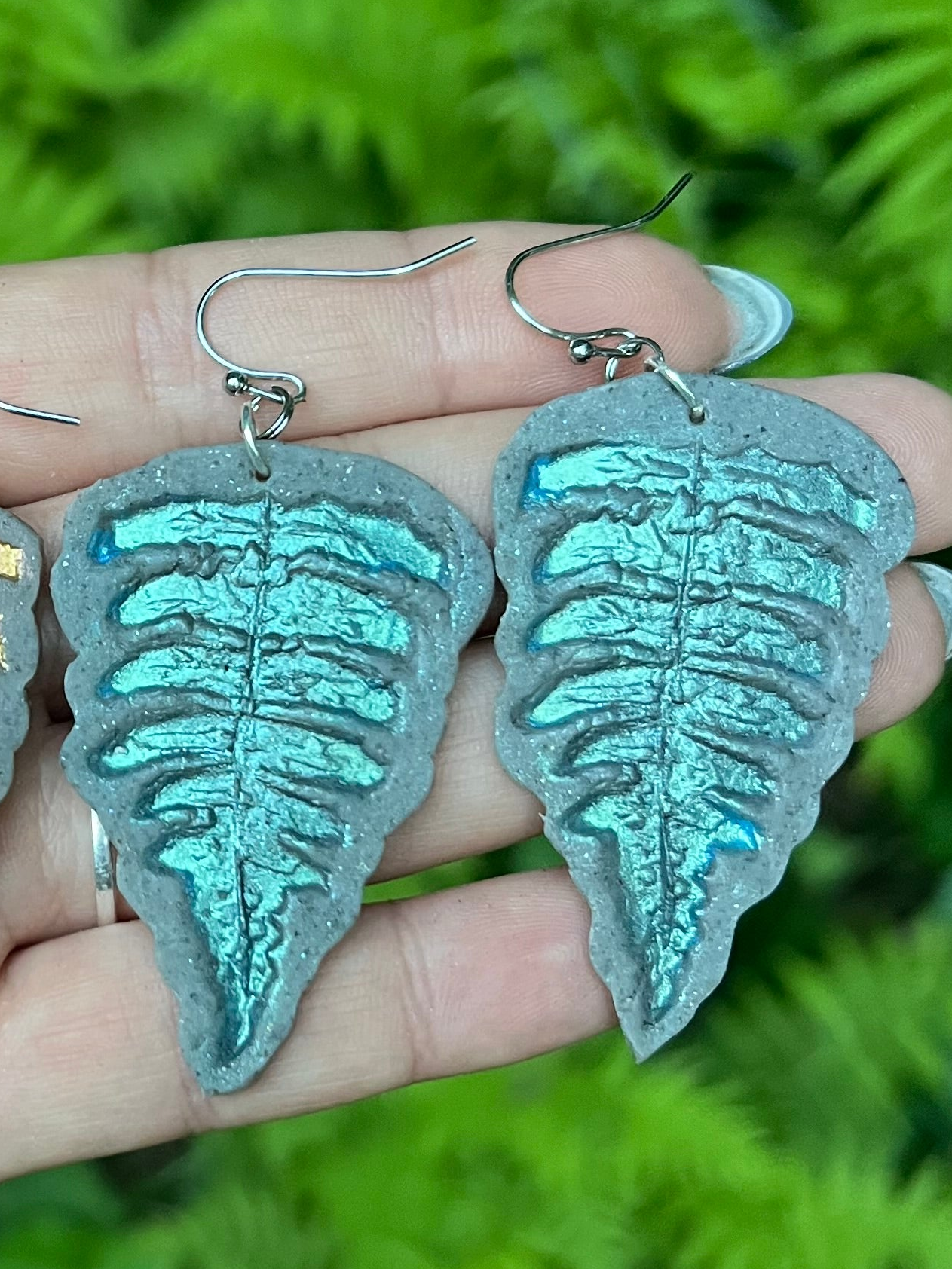 Teal fern-shaped polymer clay earrings held in a hand with a green foliage background