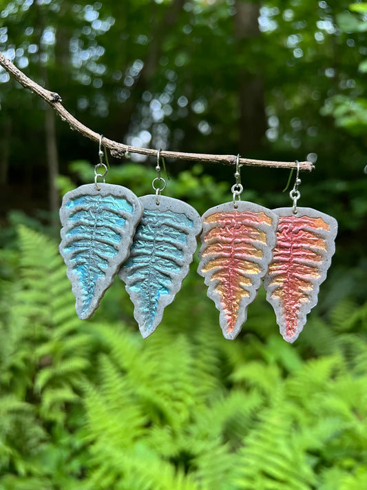 Colorful fern-shaped polymer clay earrings on a branch with a natural fern background