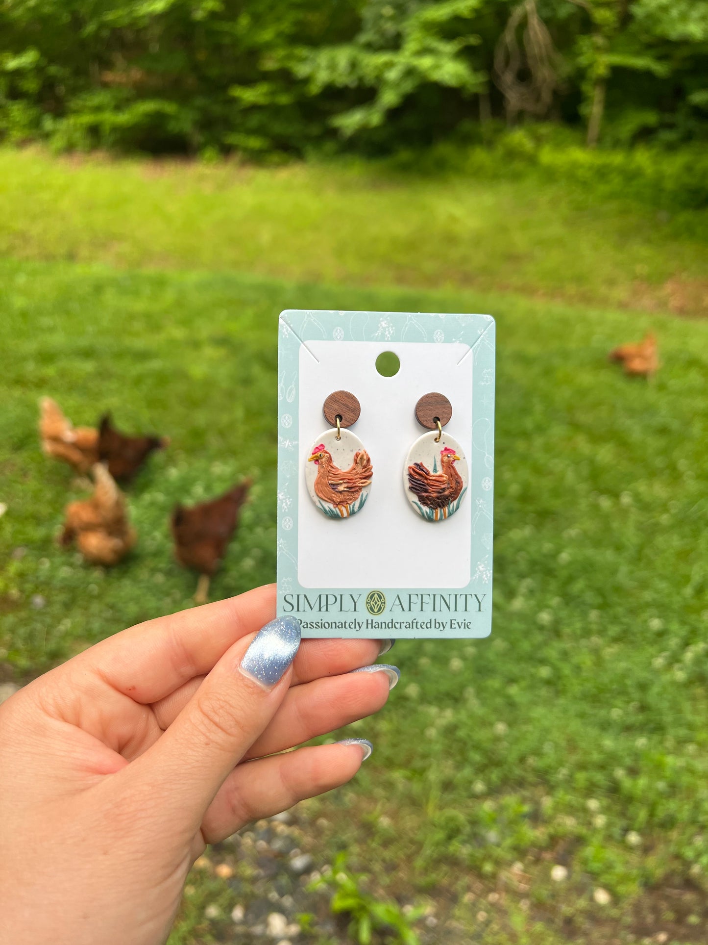 Chicken-themed polymer clay earrings on a card held in front of chickens in a grassy field
