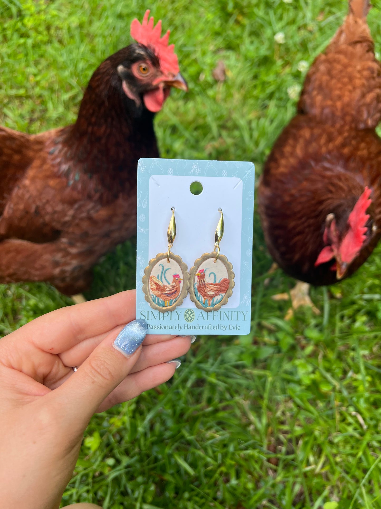 Hand holding a card with polymer clay chicken earrings in front of two chickens on grass