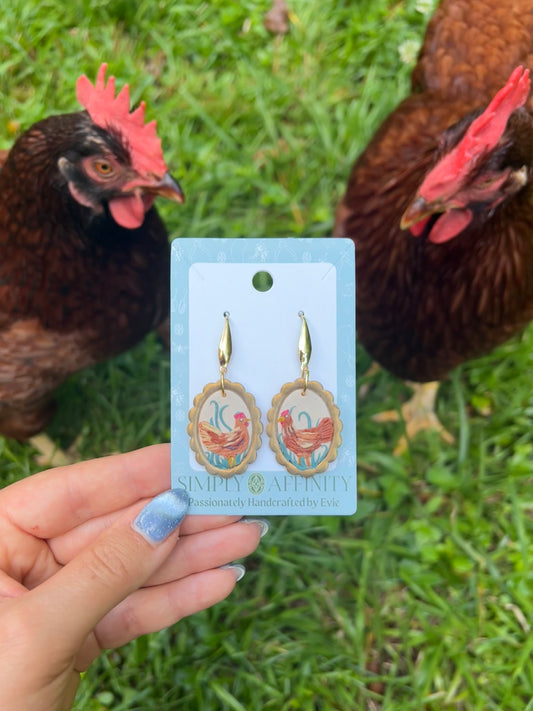 Hand holding a card with earrings featuring chicken designs, with two chickens in the background.