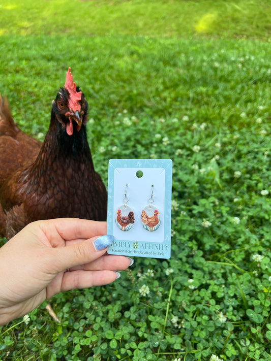 Chicken-themed polymer clay earrings on a card held in front of chickens in a grassy field