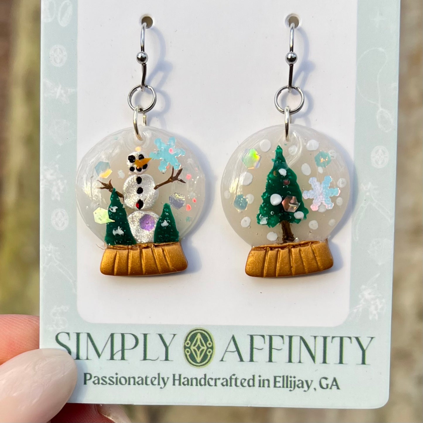 Hand sculpted polymer clay Snow globe earrings on a 'Simply Affinity' card with a blurred background