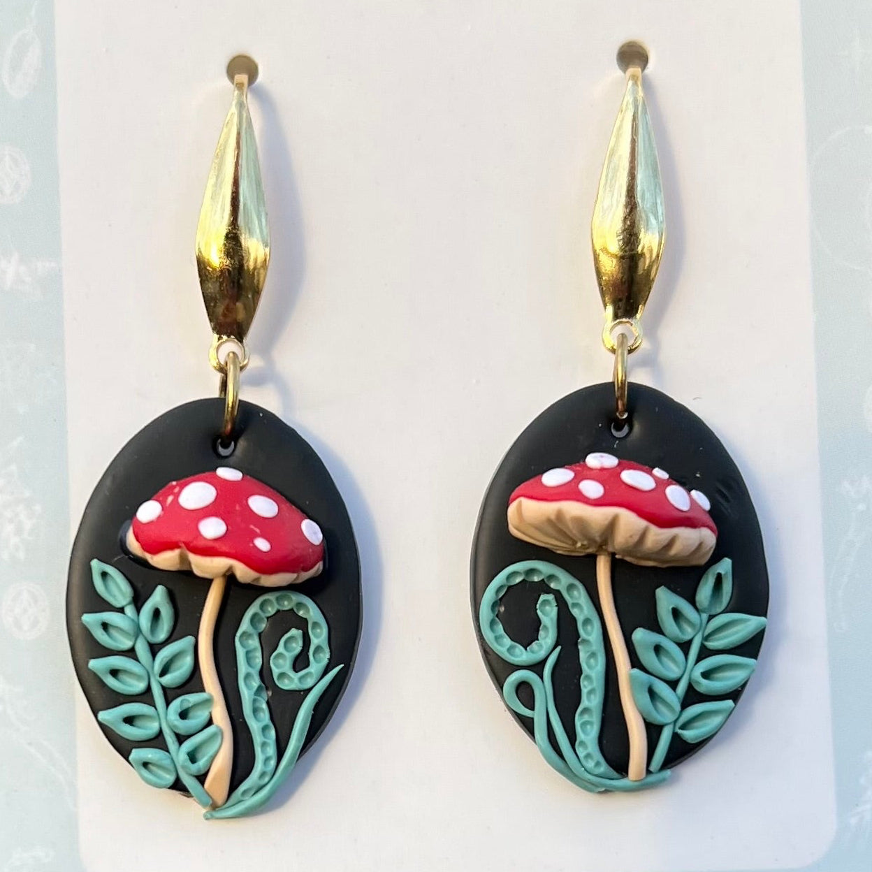 Hand sculpted polymer clay Mushroom-shaped earrings with red caps and white spots on a black oval background, featuring gold accents.