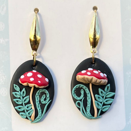 Hand sculpted polymer clay Mushroom-shaped earrings with red caps and white spots on a black oval background, featuring gold accents.