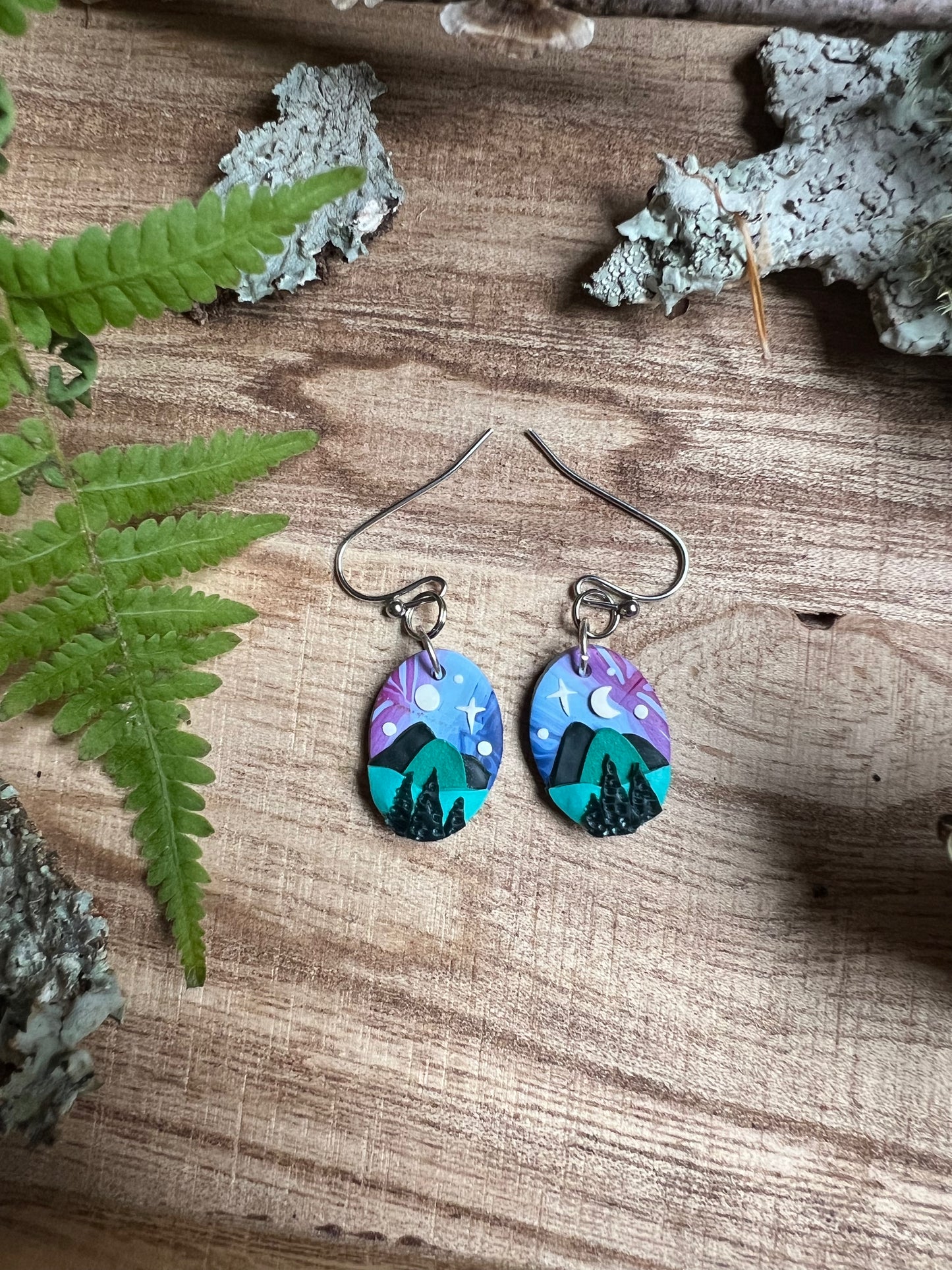 Pair of earrings with mountain design on a wooden surface with fern leaves