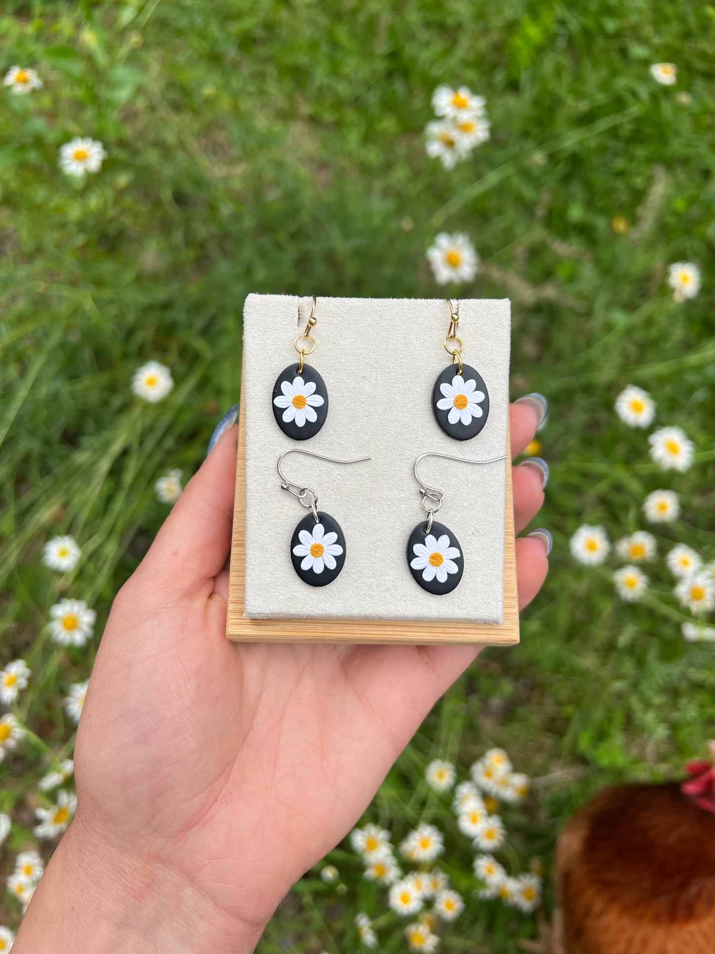 Hand holding a display card with fdaisy earrings against a natural background of grass and flowers.
