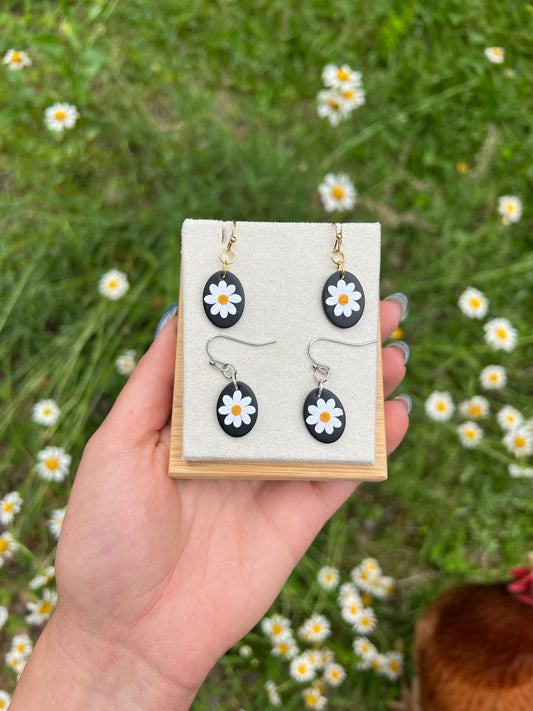 Hand holding a display card with fdaisy earrings against a natural background of grass and flowers.
