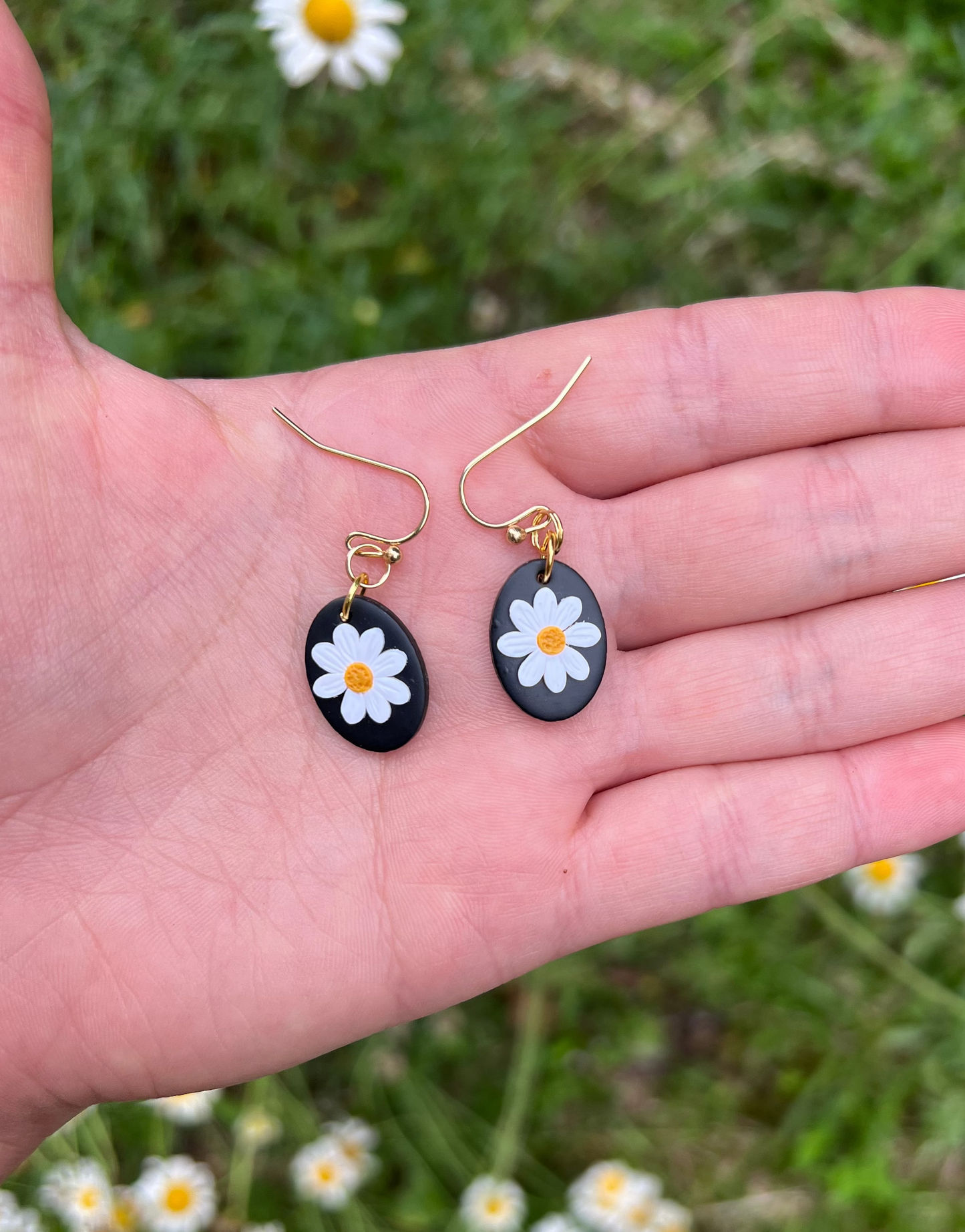 Hand holding two black and white floral daisy earrings with a natural background of greenery and flowers.