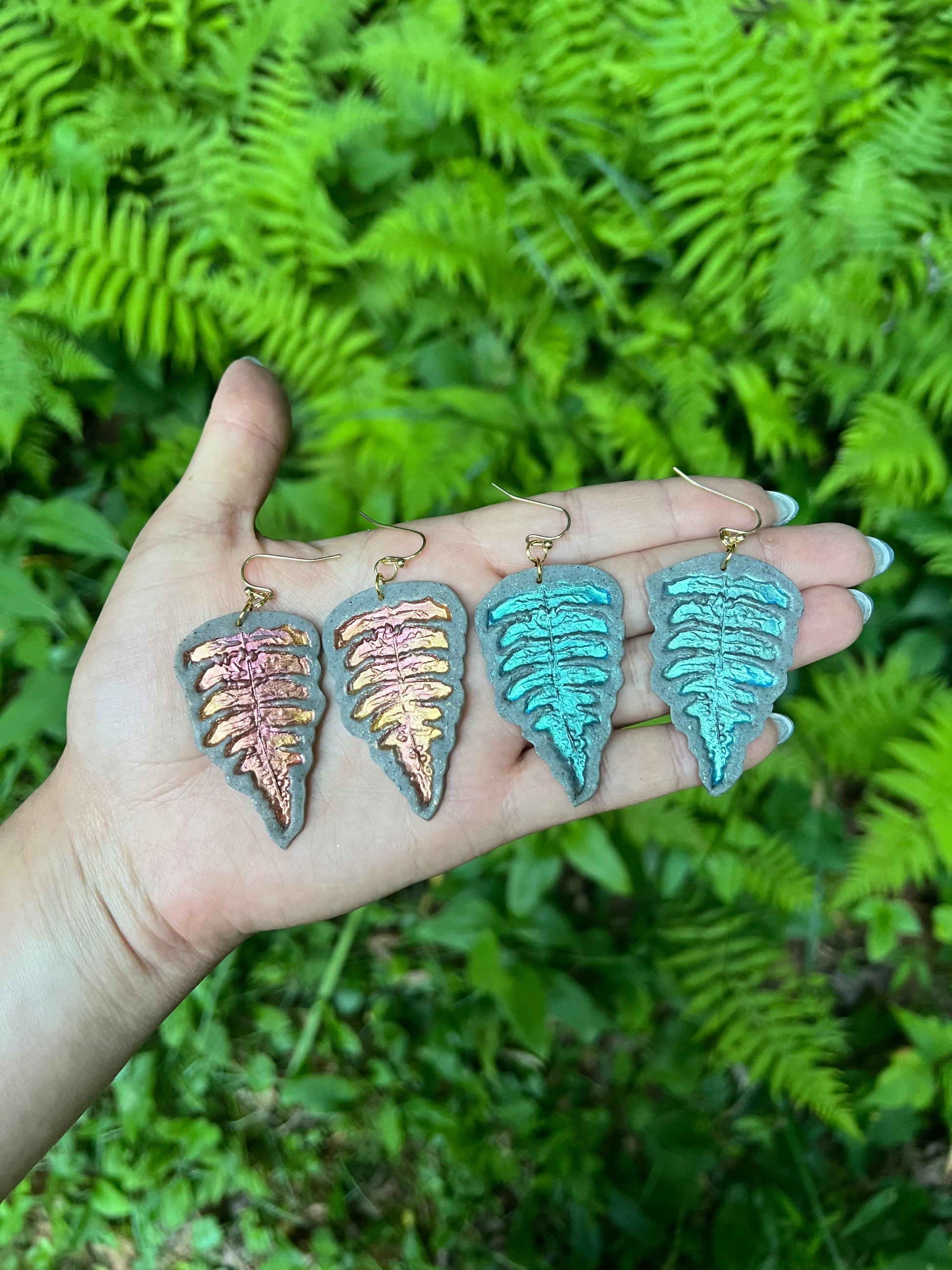 Teal  gold and copper fern-shaped polymer clay earrings held in a hand with a green foliage background