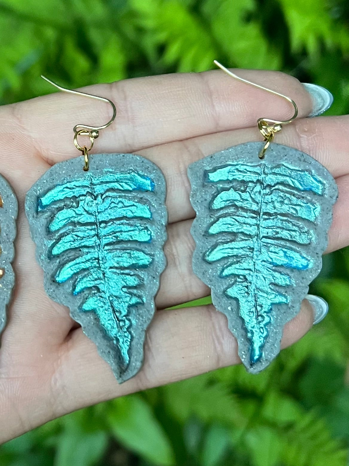 Fern leaf-shaped polymer clay earrings with turquoise metallic finish held in hand against a green leafy background