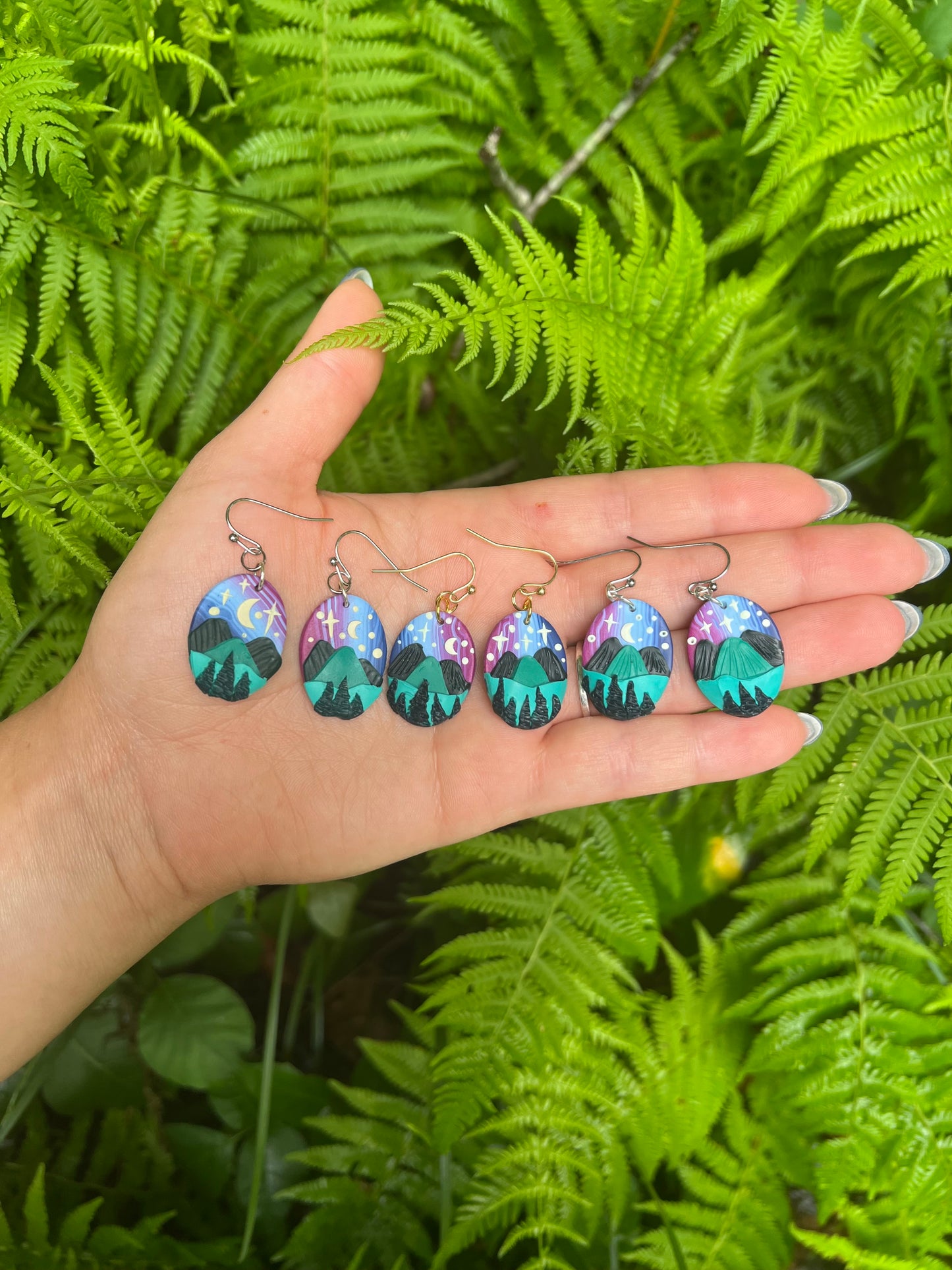 Hand holding six earrings with nature-themed designs against a green fern background