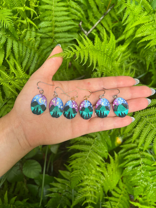 Hand holding six earrings with nature-themed designs against a green fern background