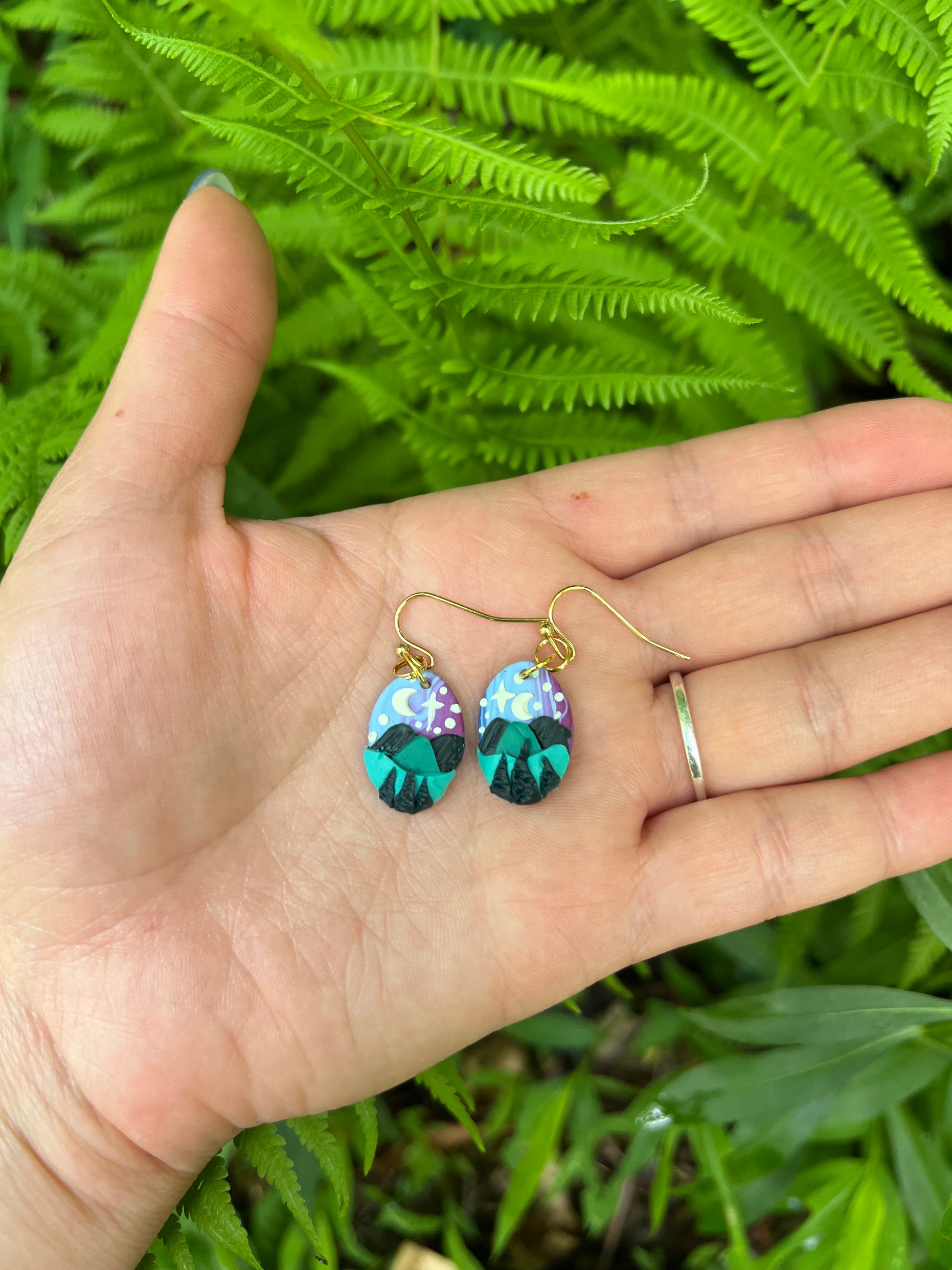 Hand holding Hand-sculpted starry blue ridge mountain polymer clay earrings on natural surface. 