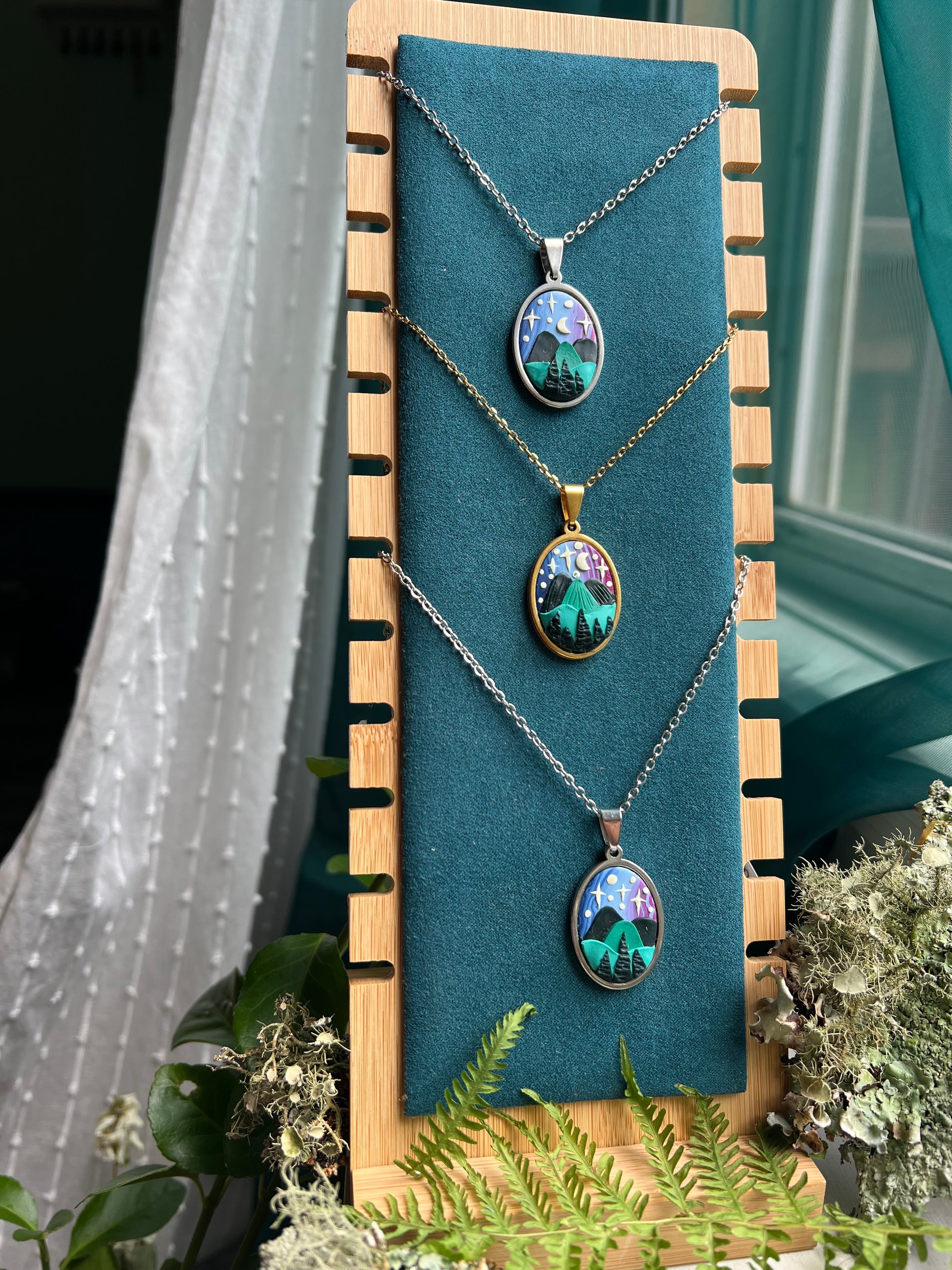 Three hand-sculpted starry blue ridge mountain polymer clay necklaces with nature-themed pendants on necklace stand 
