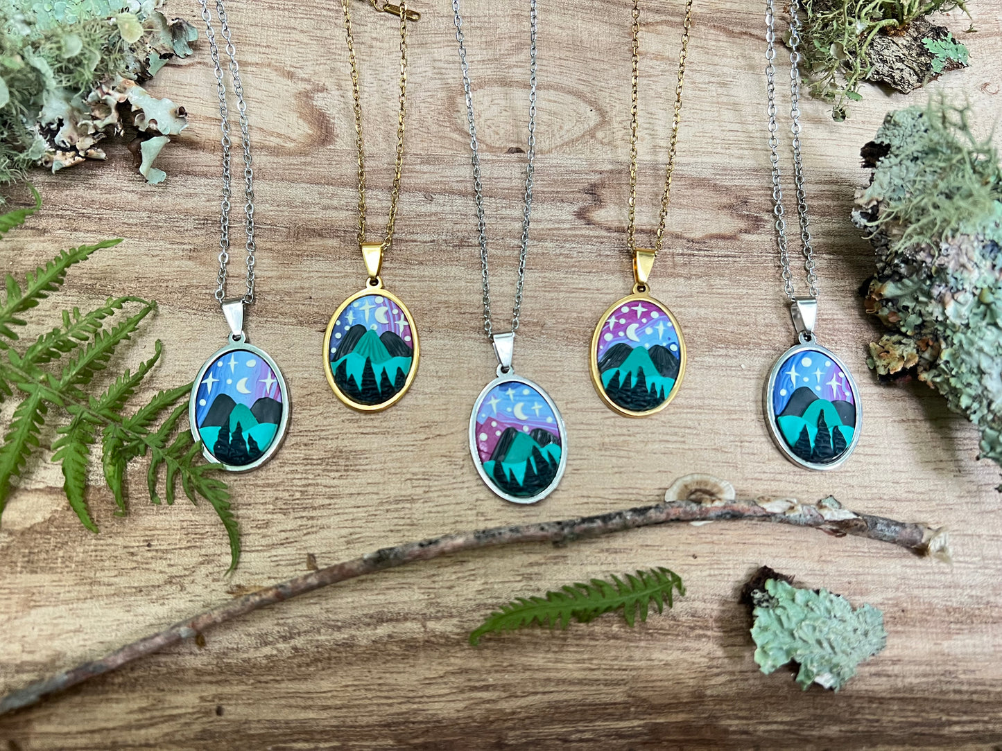 Five necklaces with nature-themed pendants on a wooden surface with greenery.