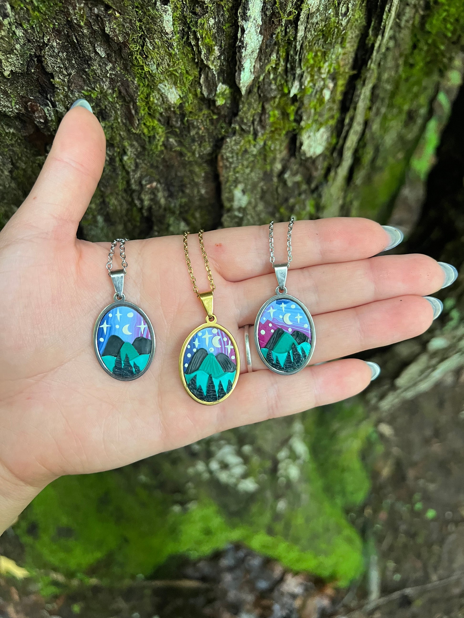 Hand holding three hand-sculpted starry blue ridge mountain polymer clay necklaces with nature-themed pendants on a mossy surface.