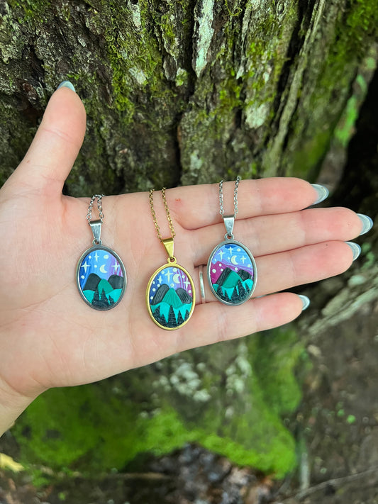 Hand holding three hand-sculpted starry blue ridge mountain polymer clay necklaces with nature-themed pendants on a mossy surface.