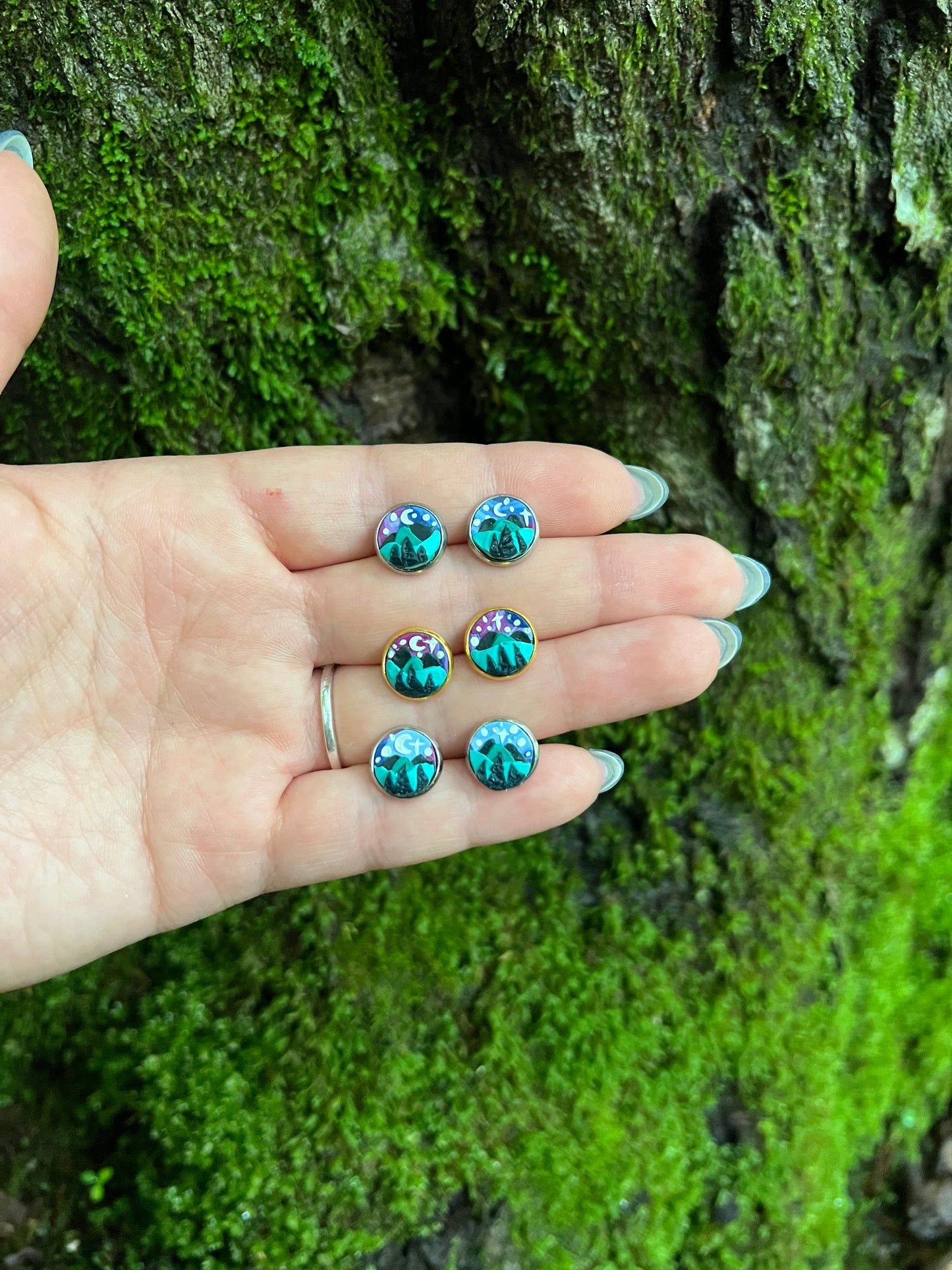 Hand holding hand sculpted polymer clay stud earrings with a starry mountain scene mossy stone background