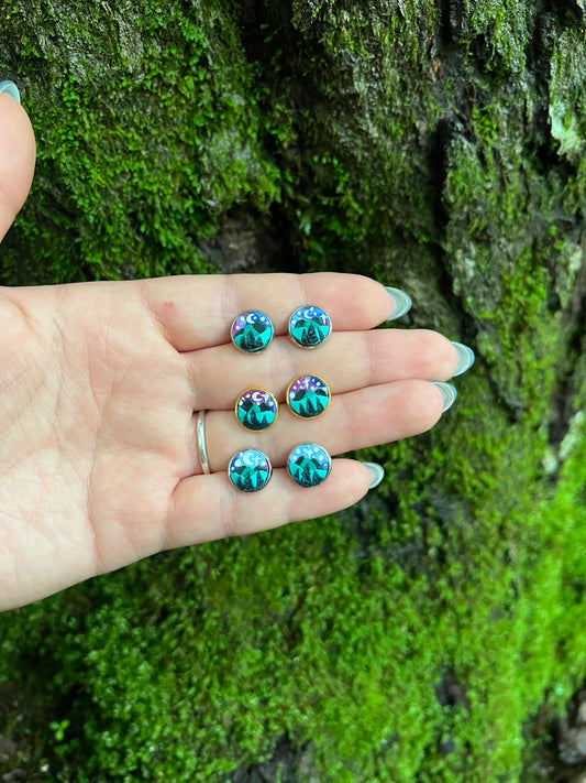 Hand holding hand sculpted polymer clay stud earrings with a starry mountain scene mossy stone background