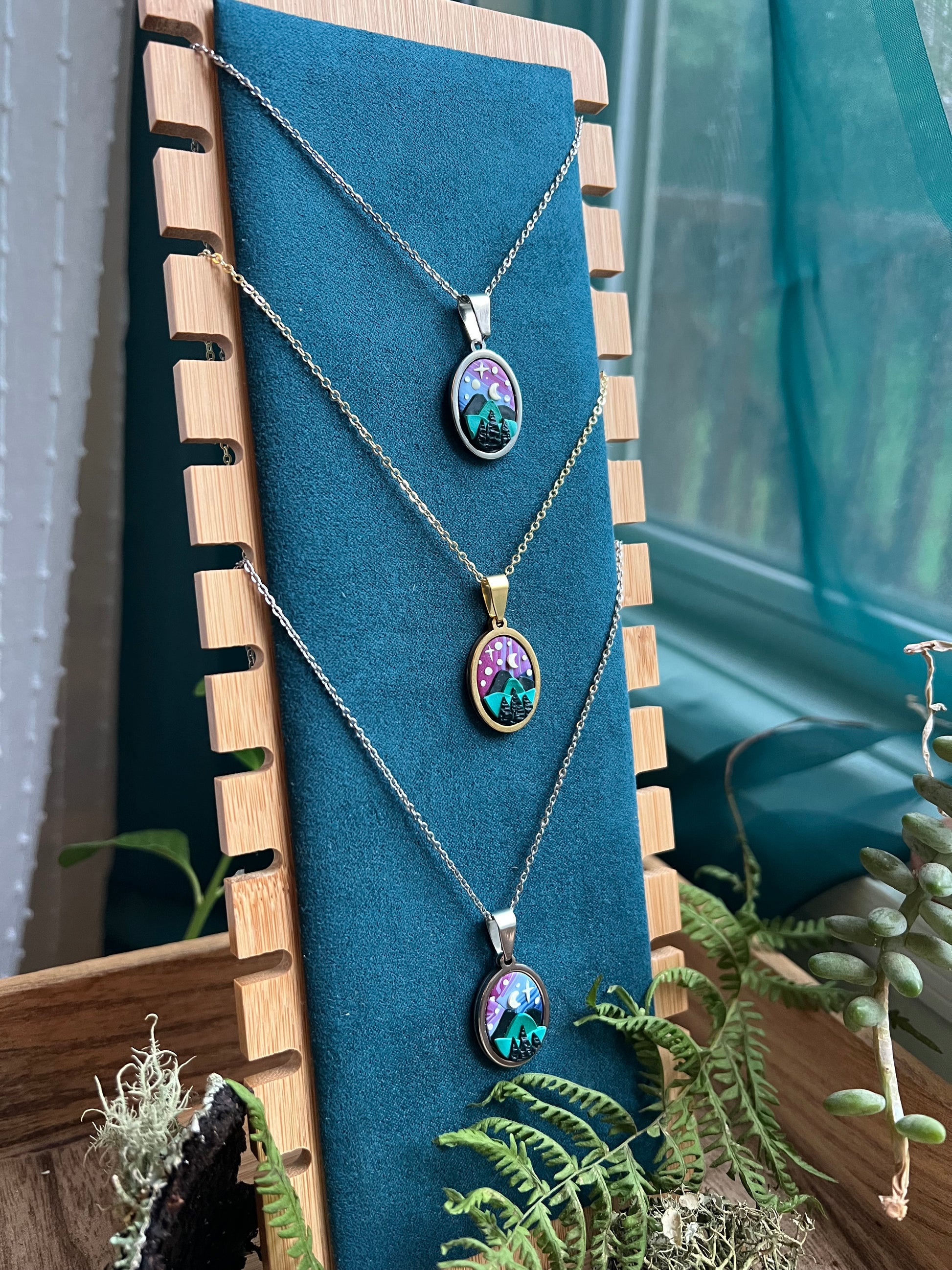 Three starry blue ridge mountain necklaces with colorful pendants on a blue cushion with a wooden border, surrounded by plants.