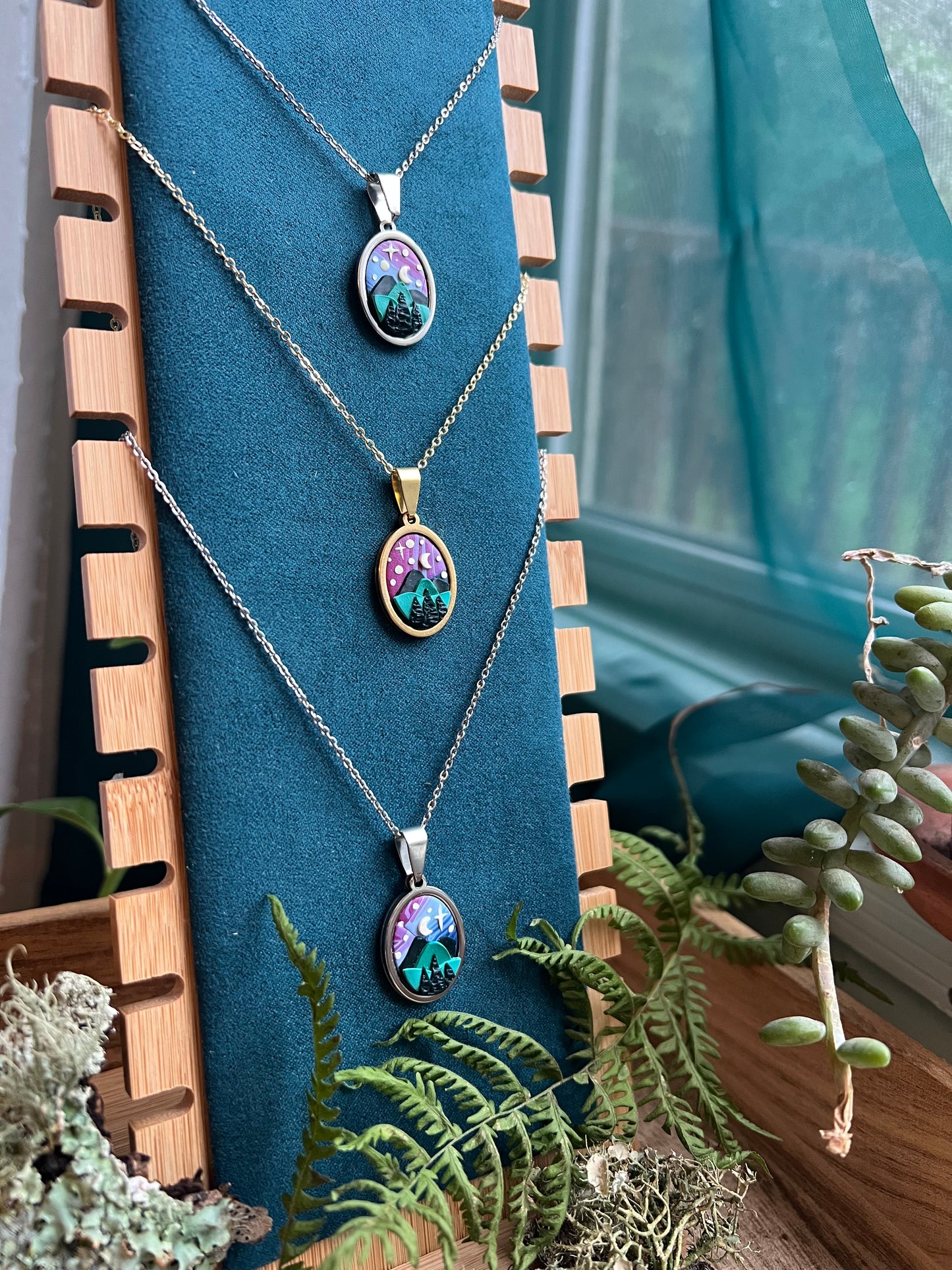 Three polymer clay necklaces with pendants on a blue stand with plants in the background