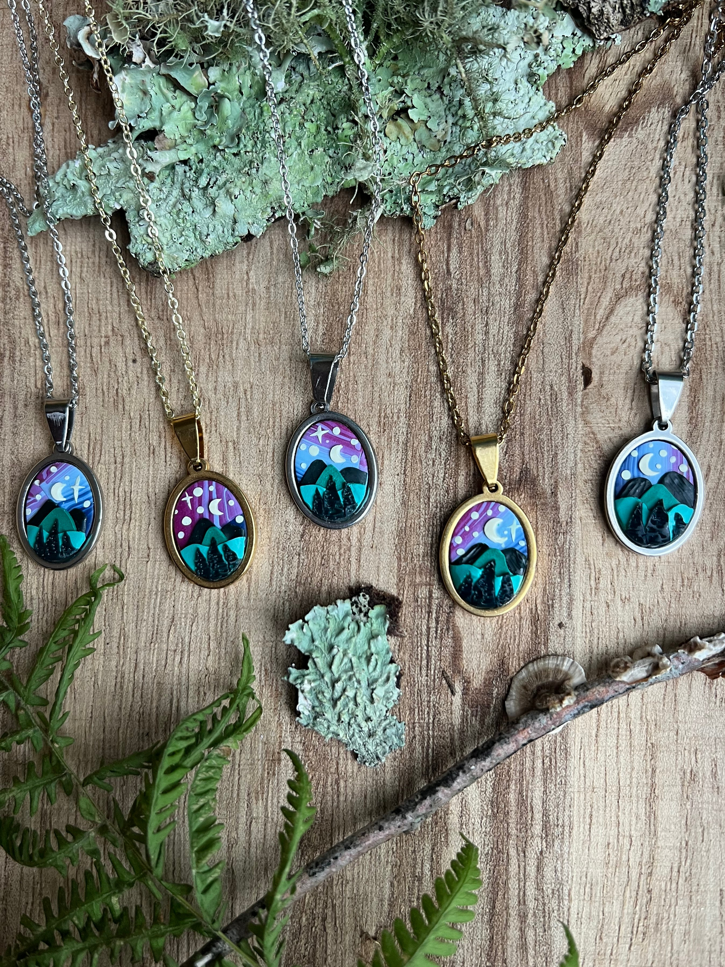 Necklaces with nature-themed starry mountain pendants on a wooden surface with moss and leaves.