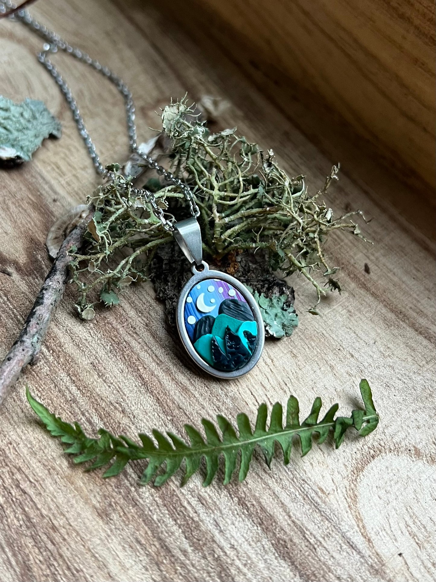 Necklace with a pendant featuring a night sky design, surrounded by greenery on a wooden surface.