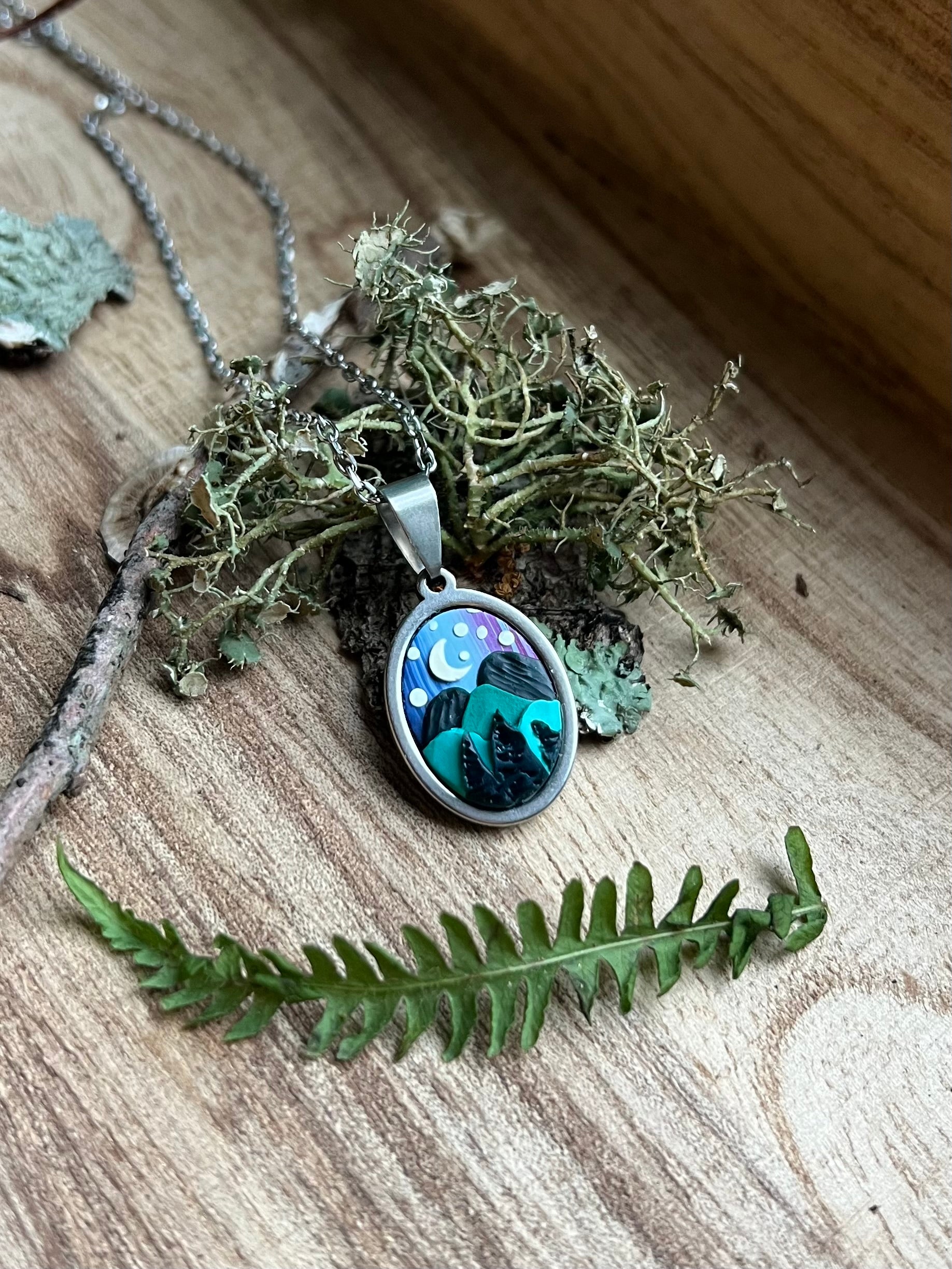 Necklace with a pendant featuring a night sky design, surrounded by greenery on a wooden surface.