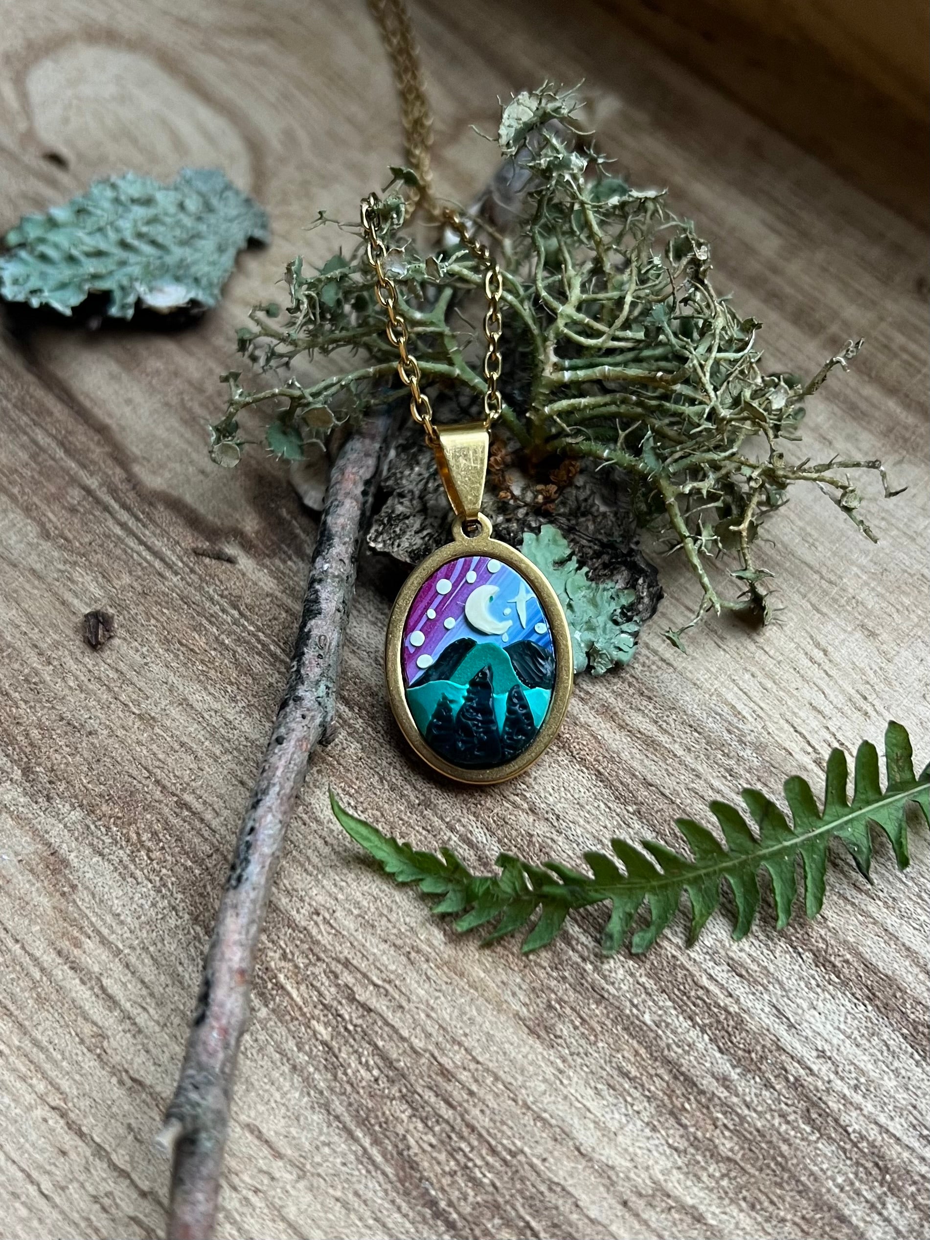 Necklace with a pendant featuring a mountain and starry sky design, placed on a wooden surface with green leaves.
