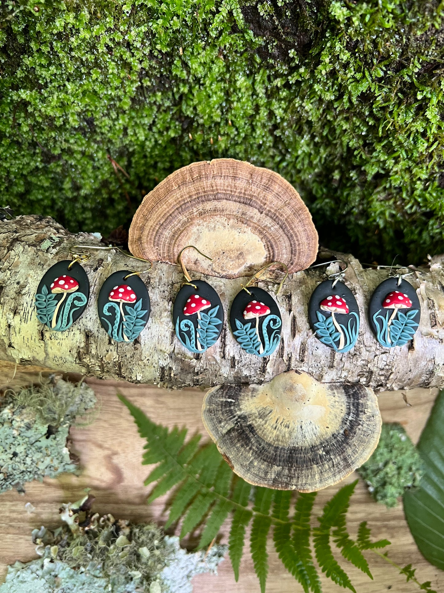 Set of five mushroom-themed earrings on a wooden log with green foliage in the background
