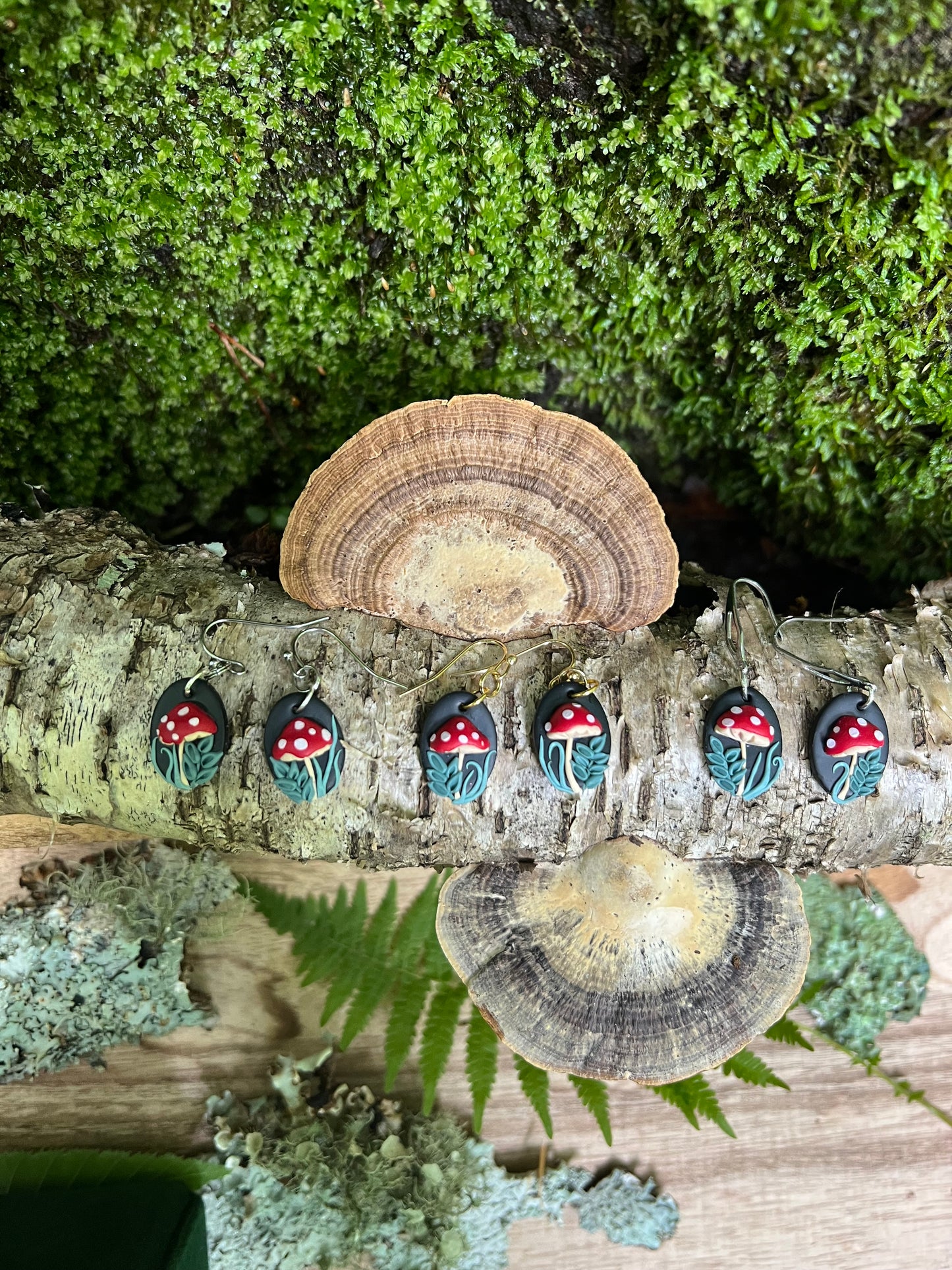 Polymer Clay Mushroom earrings on a tree trunk with mushrooms and greenery.