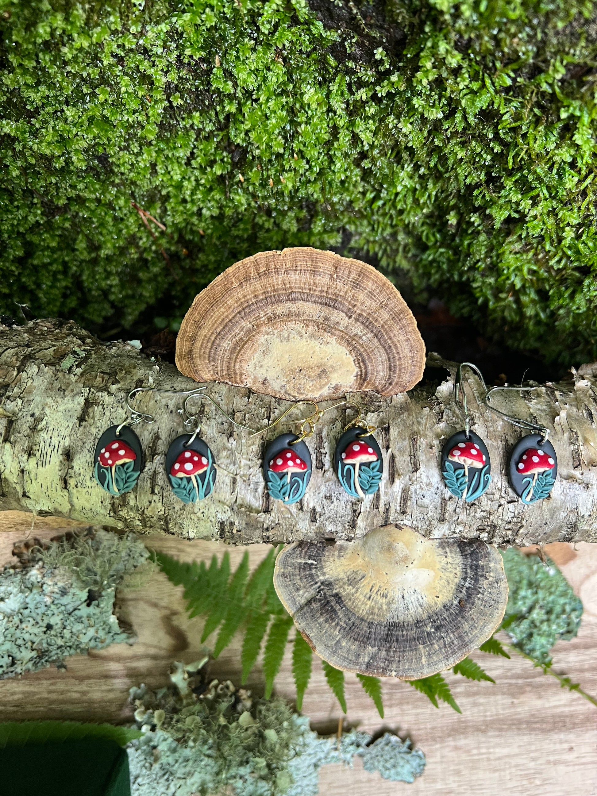 Polymer Clay Mushroom earrings on a tree trunk with mushrooms and greenery.