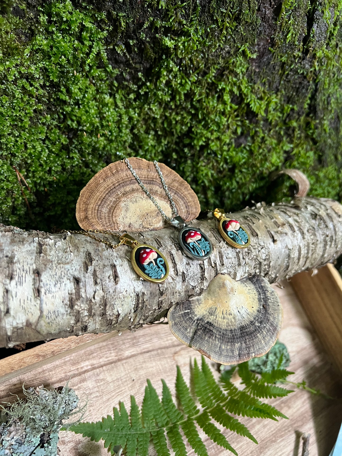 Mushroom necklaces with nature-themed pendants on a wooden log with moss and ferns.