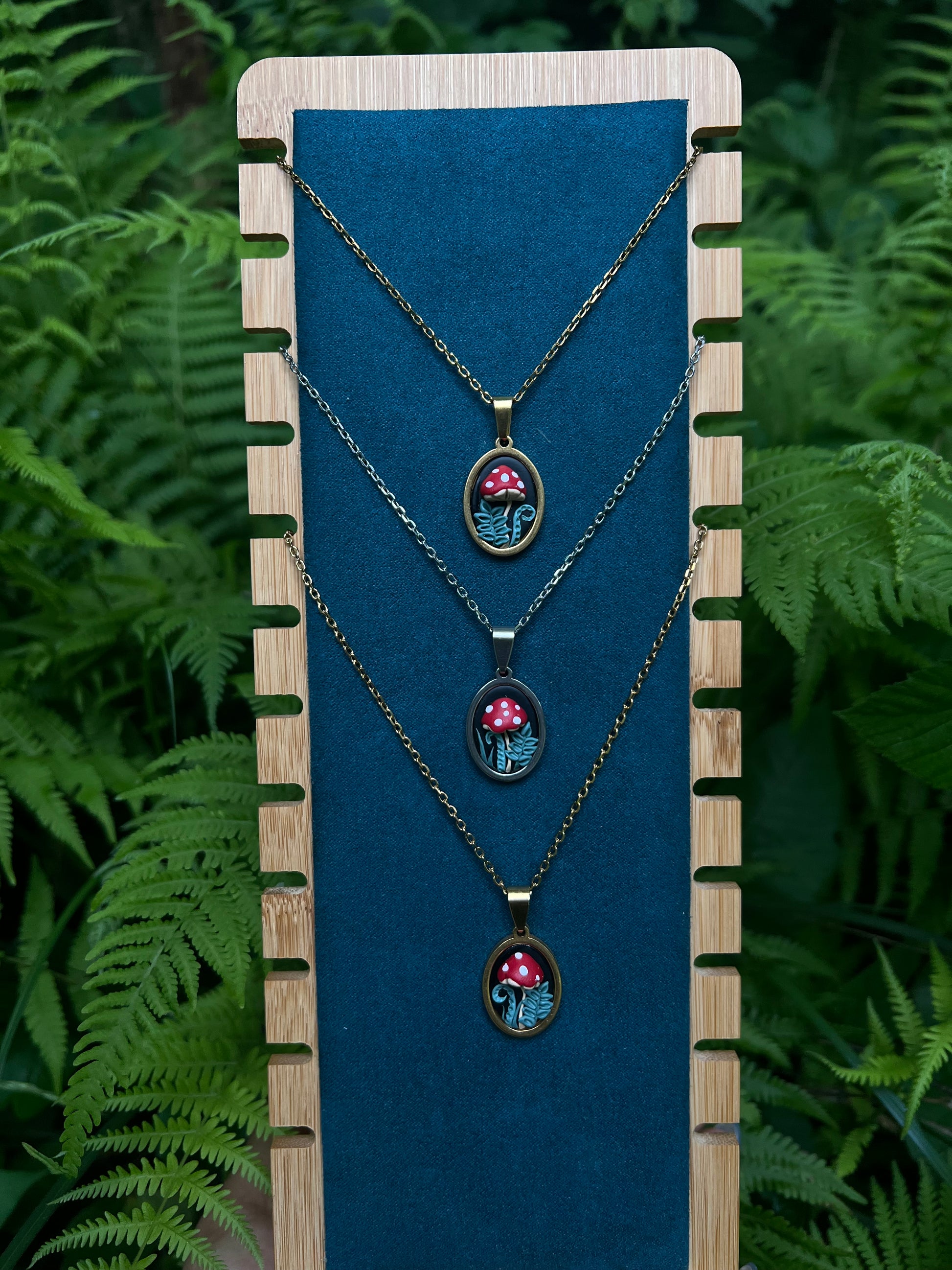 Three necklaces with mushroom pendants on a blue card against a green fern background