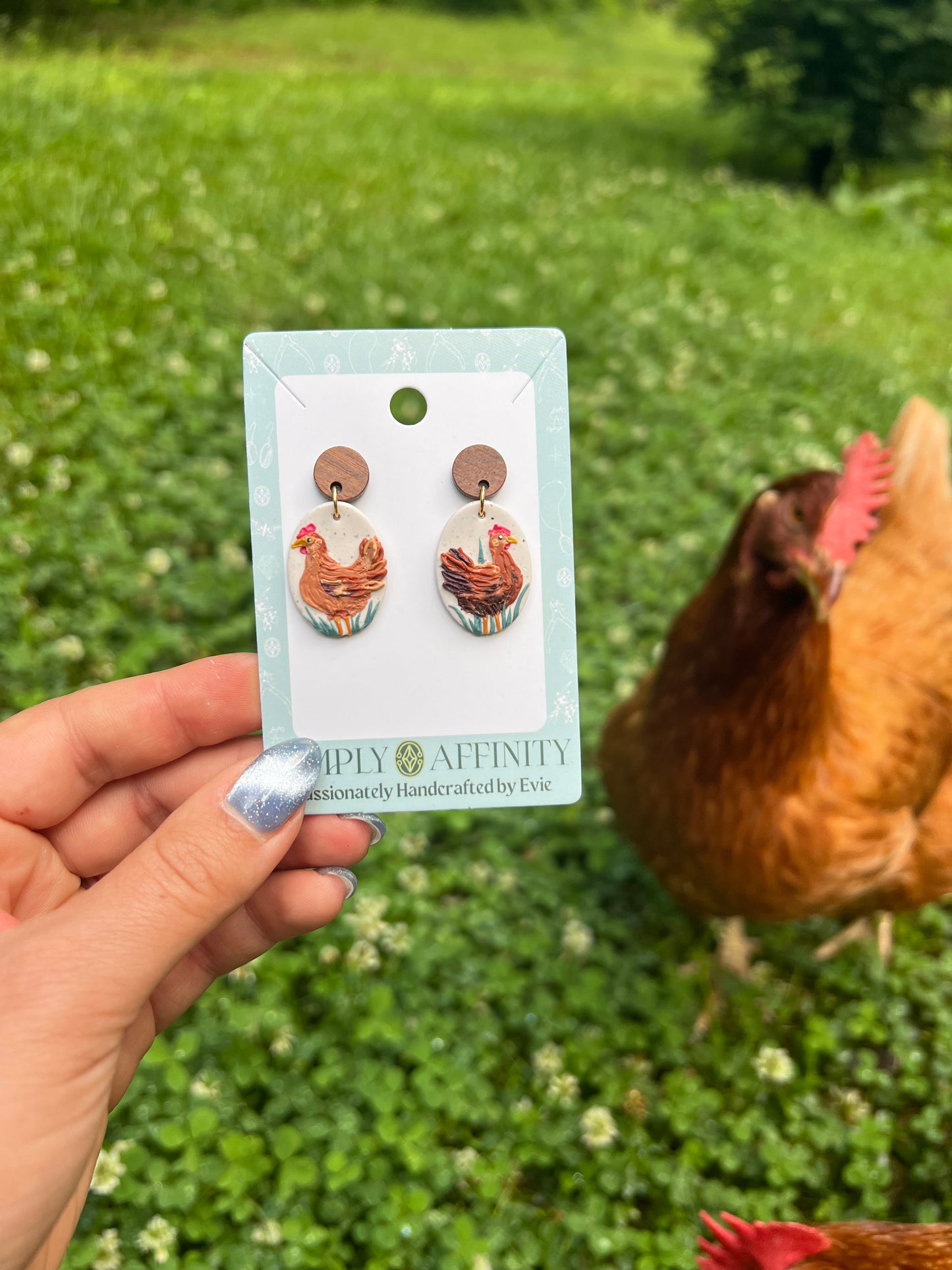Chicken-themed earrings on a card held in front of chickens in a grassy field