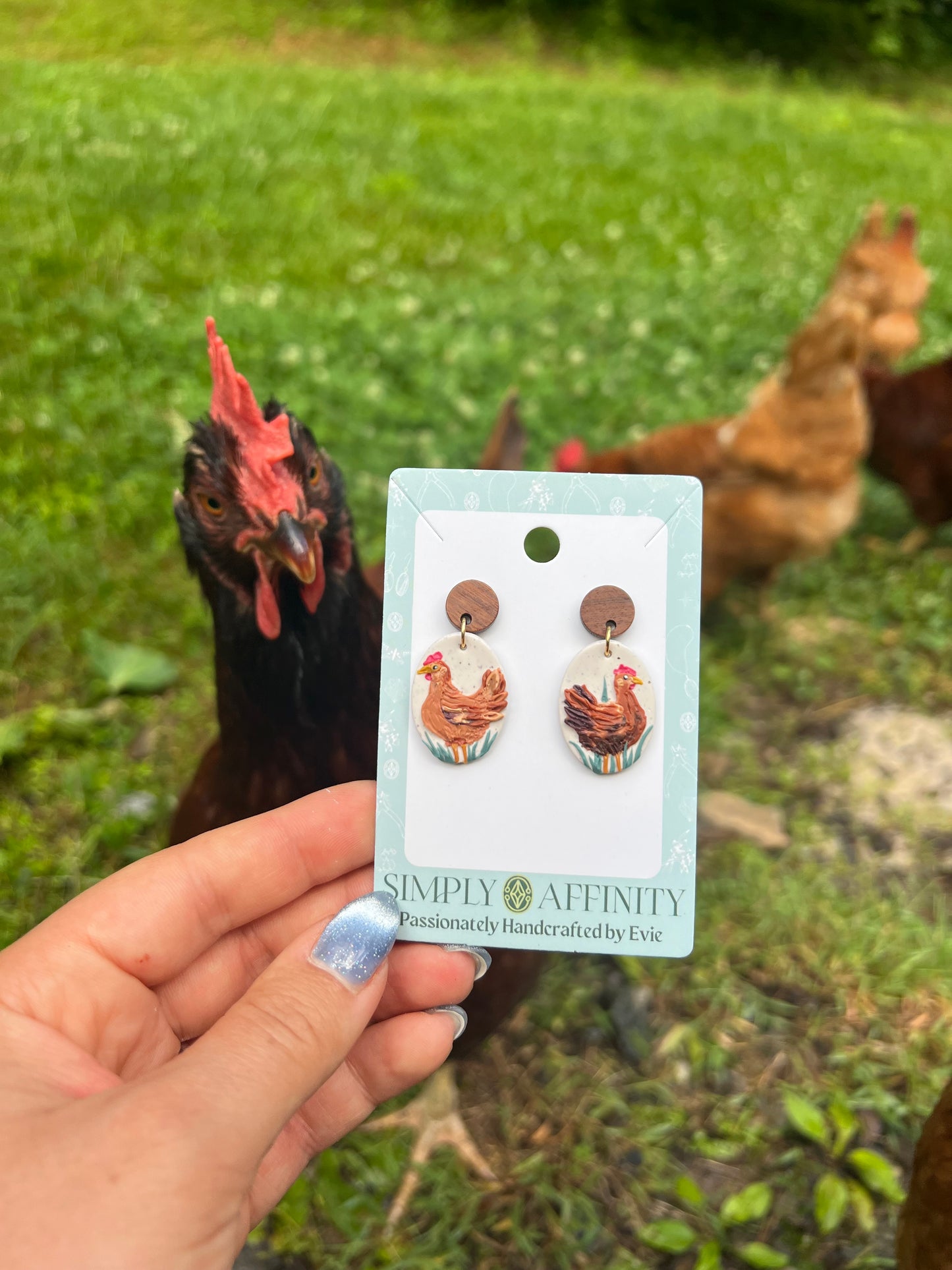 Chicken-themed polymer clay earrings on a card held in front of chickens in a grassy field