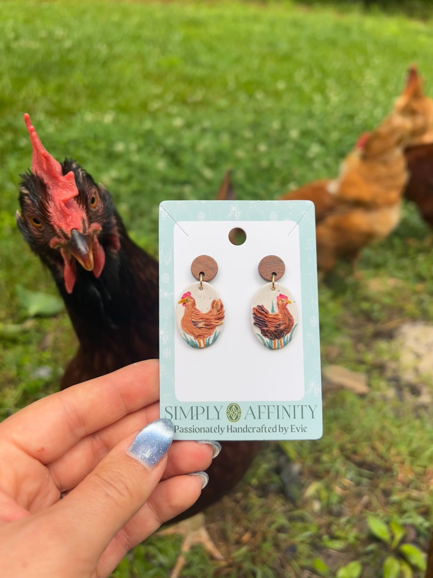 Chicken-themed earrings on a card held in front of chickens in a grassy field