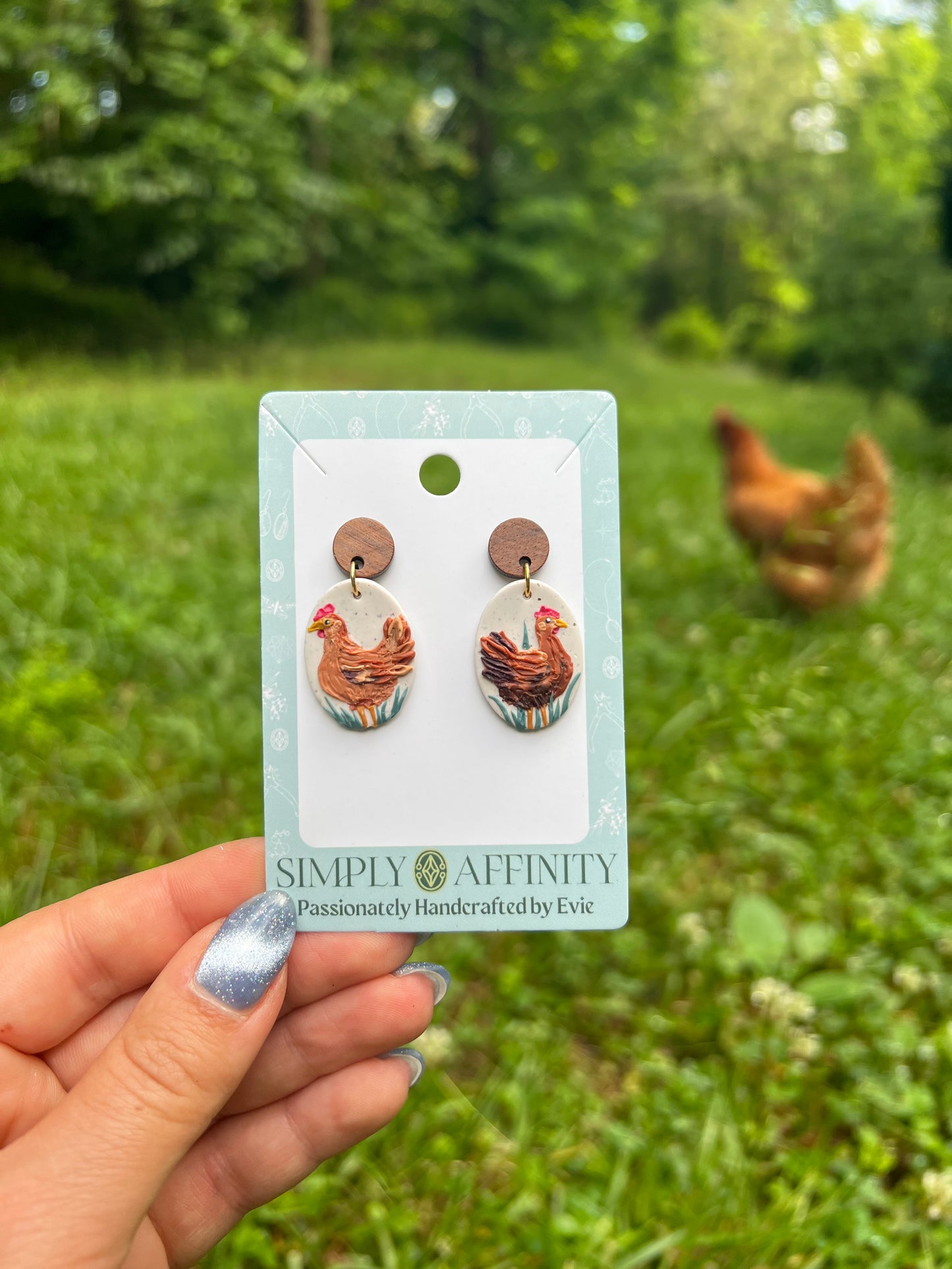 Chicken-themed polymer clay earrings on a card held in front of chickens in a grassy field