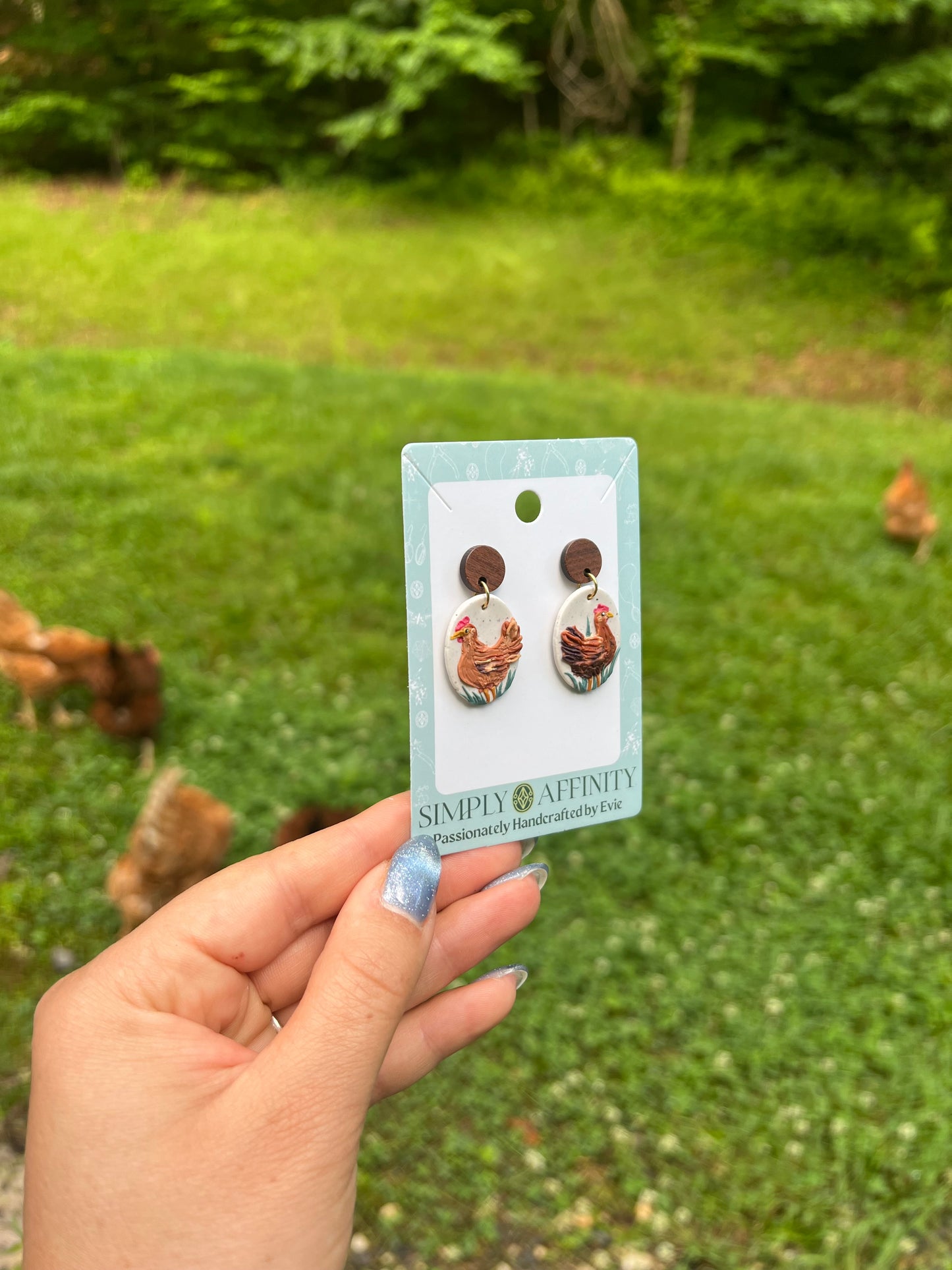 Chicken-themed earrings on a card held in front of chickens in a grassy field