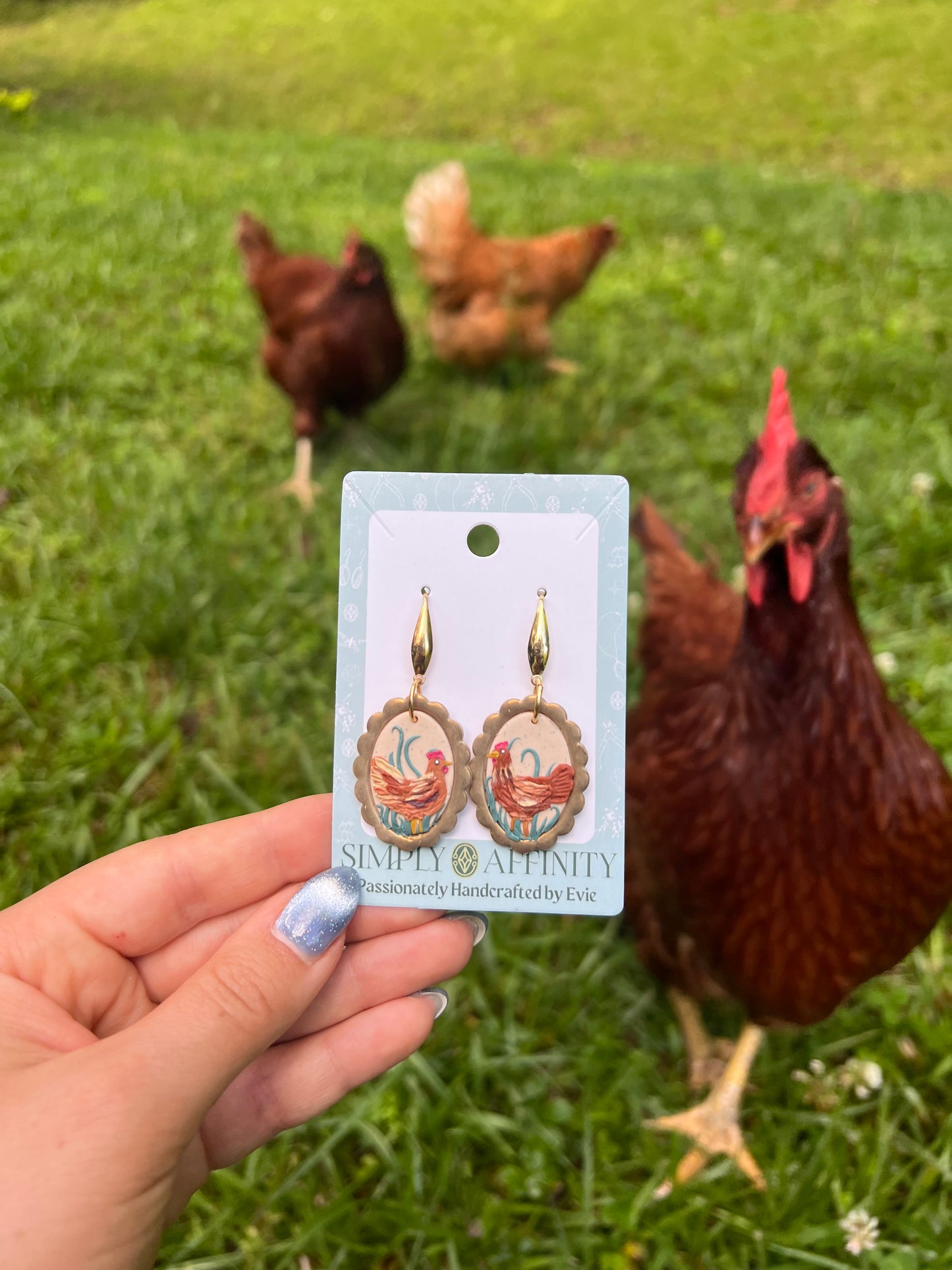 Earring card with chicken design held in front of chickens on grass