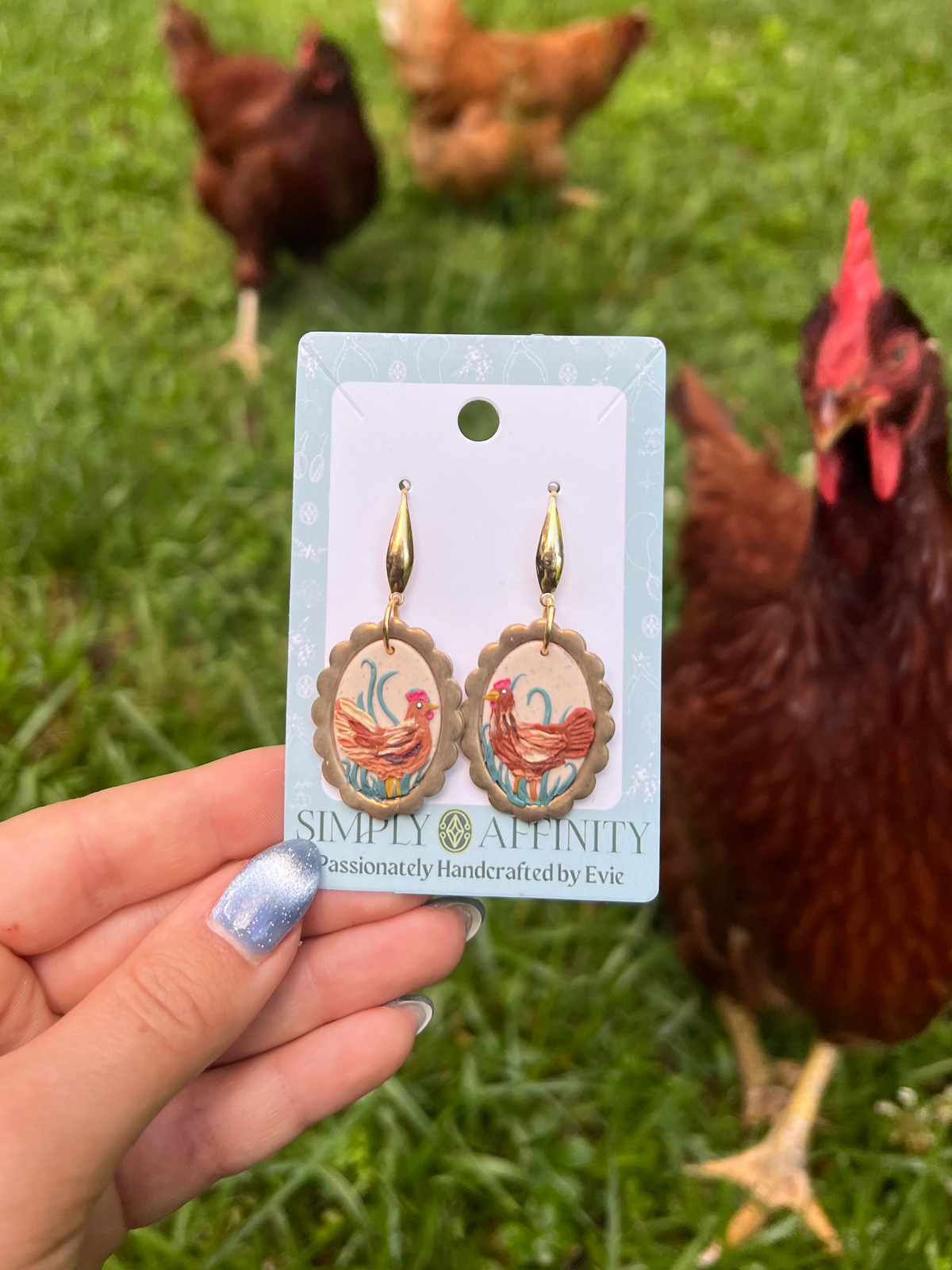 Chicken-themed earrings on a card held by a hand with chickens in the background
