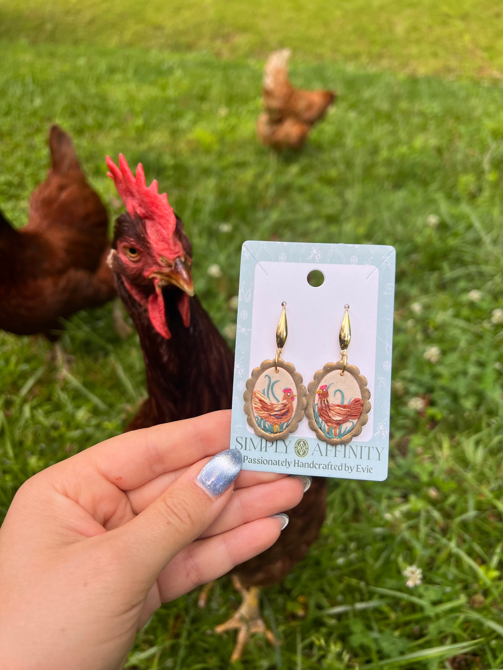 Hand holding a card with earrings featuring chicken designs, with hens in the background.