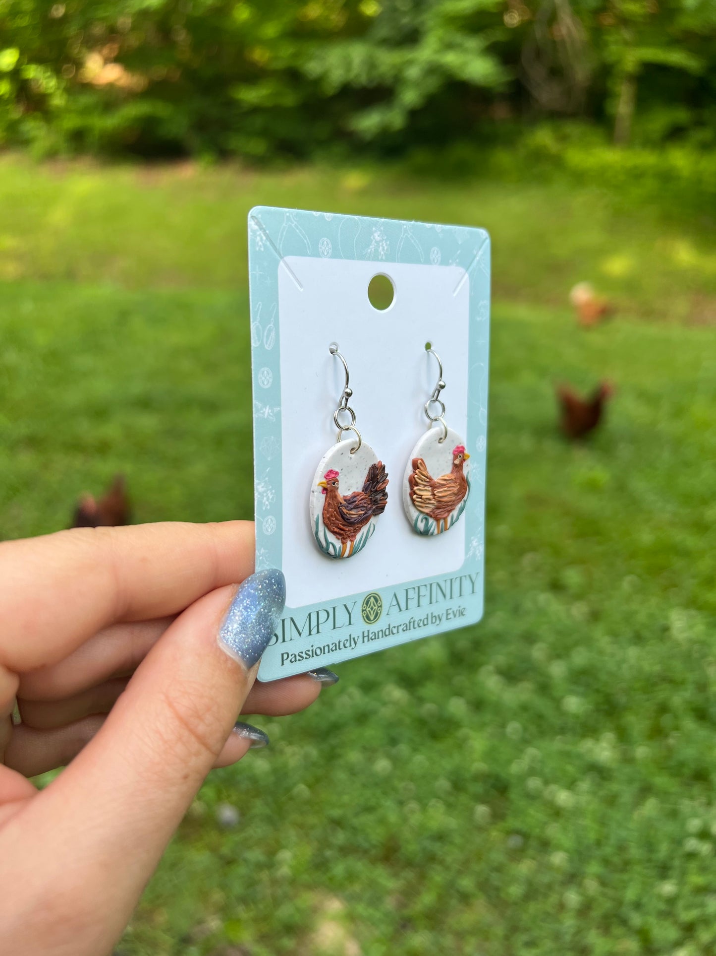 Chicken-themed polymer clay earrings on a card held in front of chickens in a grassy field