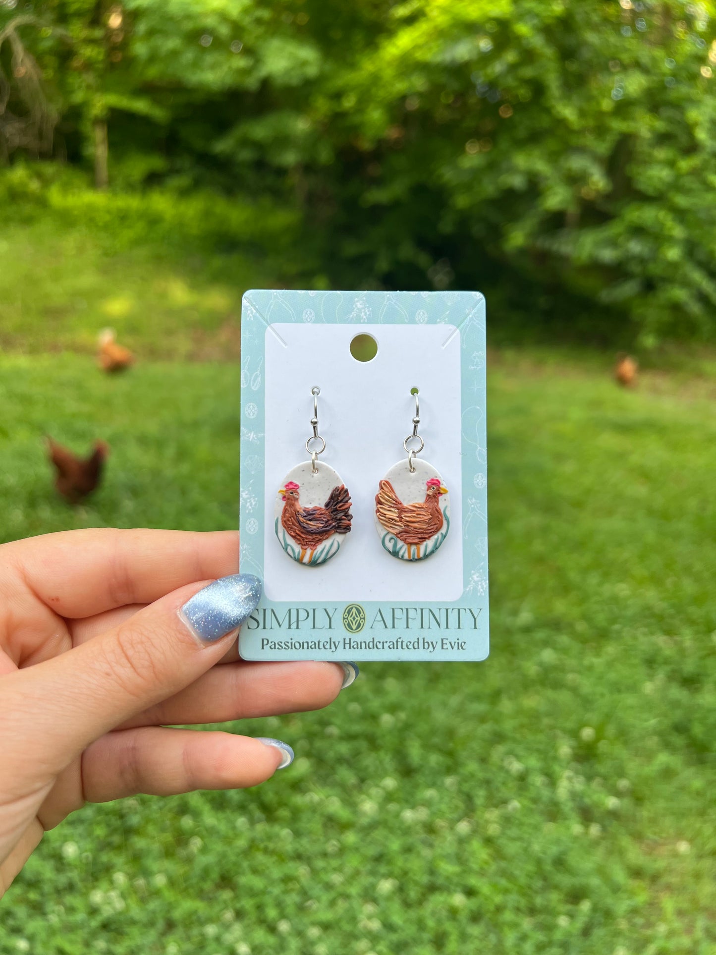 Chicken-themed polymer clay earrings on a card held in front of chickens in a grassy field