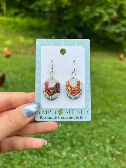 Chicken-themed polymer clay earrings on a card held in front of chickens in a grassy field