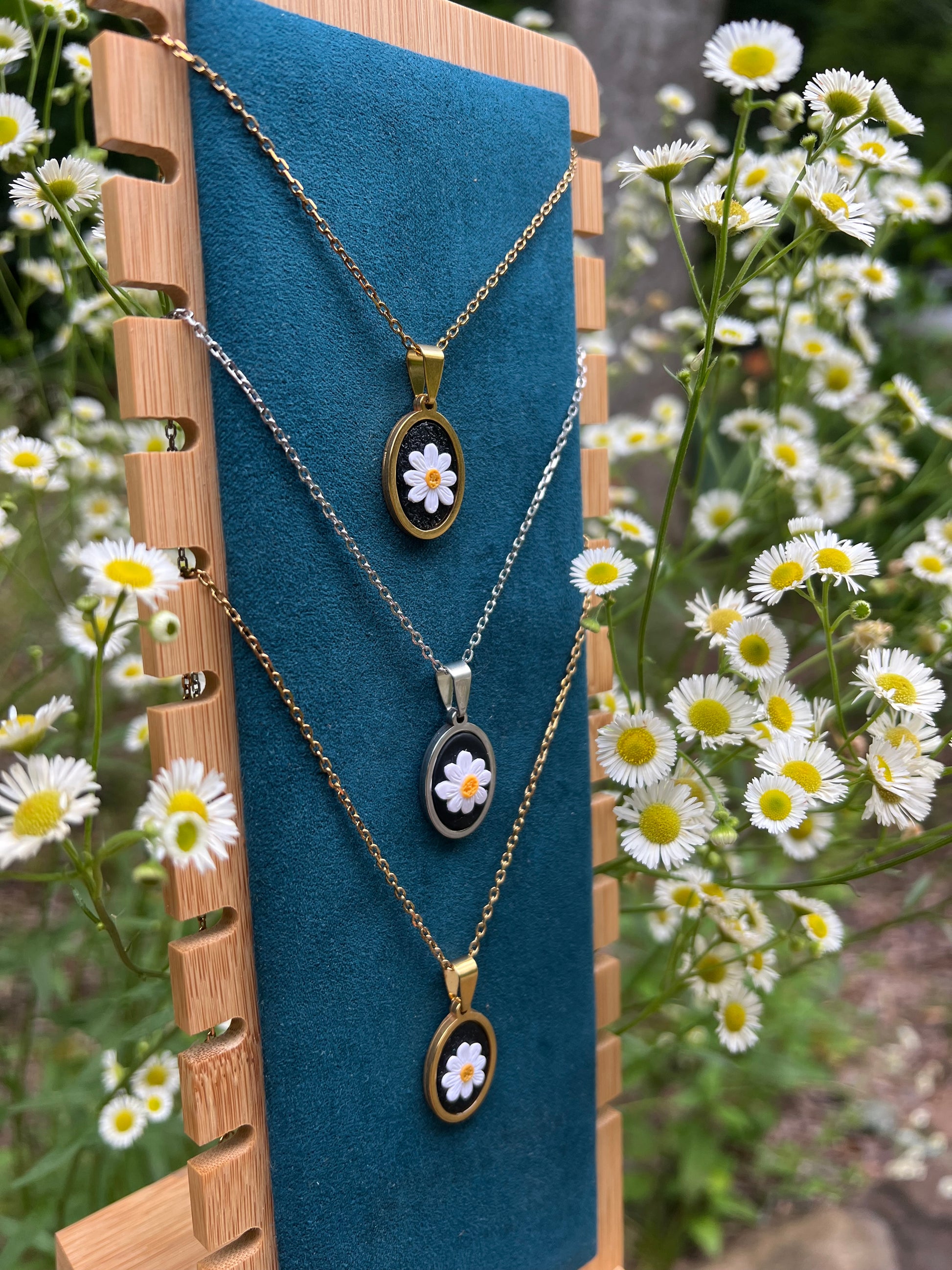 Three necklaces with daisy pendants on a blue stand, surrounded by daisies.