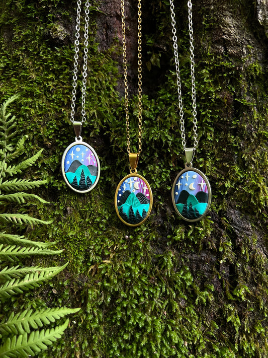 Three hand-sculpted starry blue ridge mountain polymer clay necklaces with nature-themed pendants on a mossy surface.